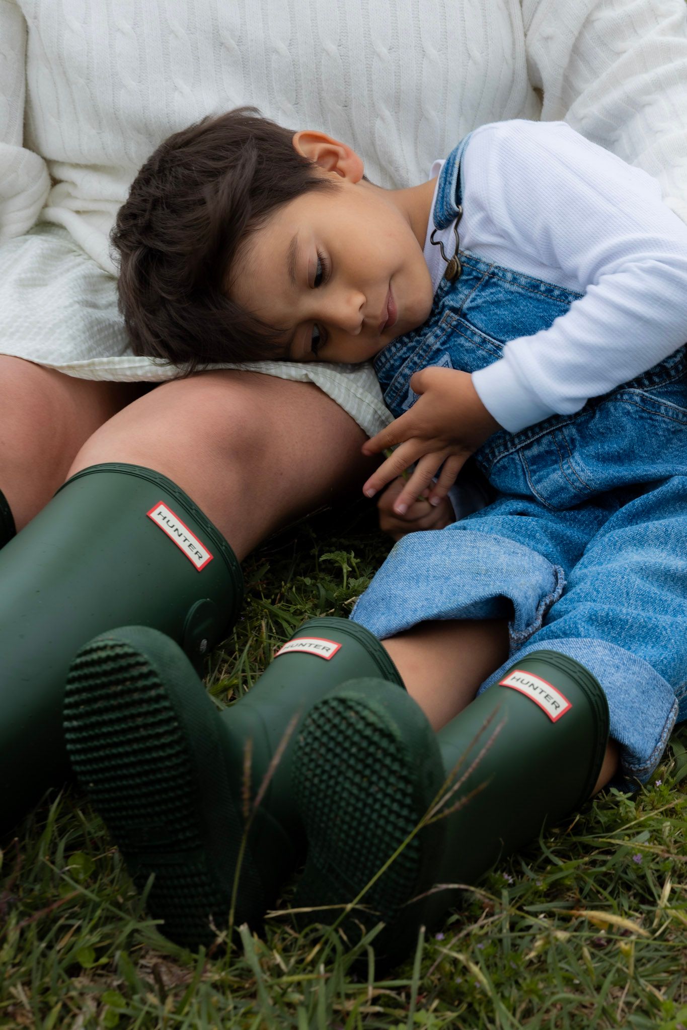 A young boy in denim overalls and green Hunter boots rests his head on his mothers lap outdoors.