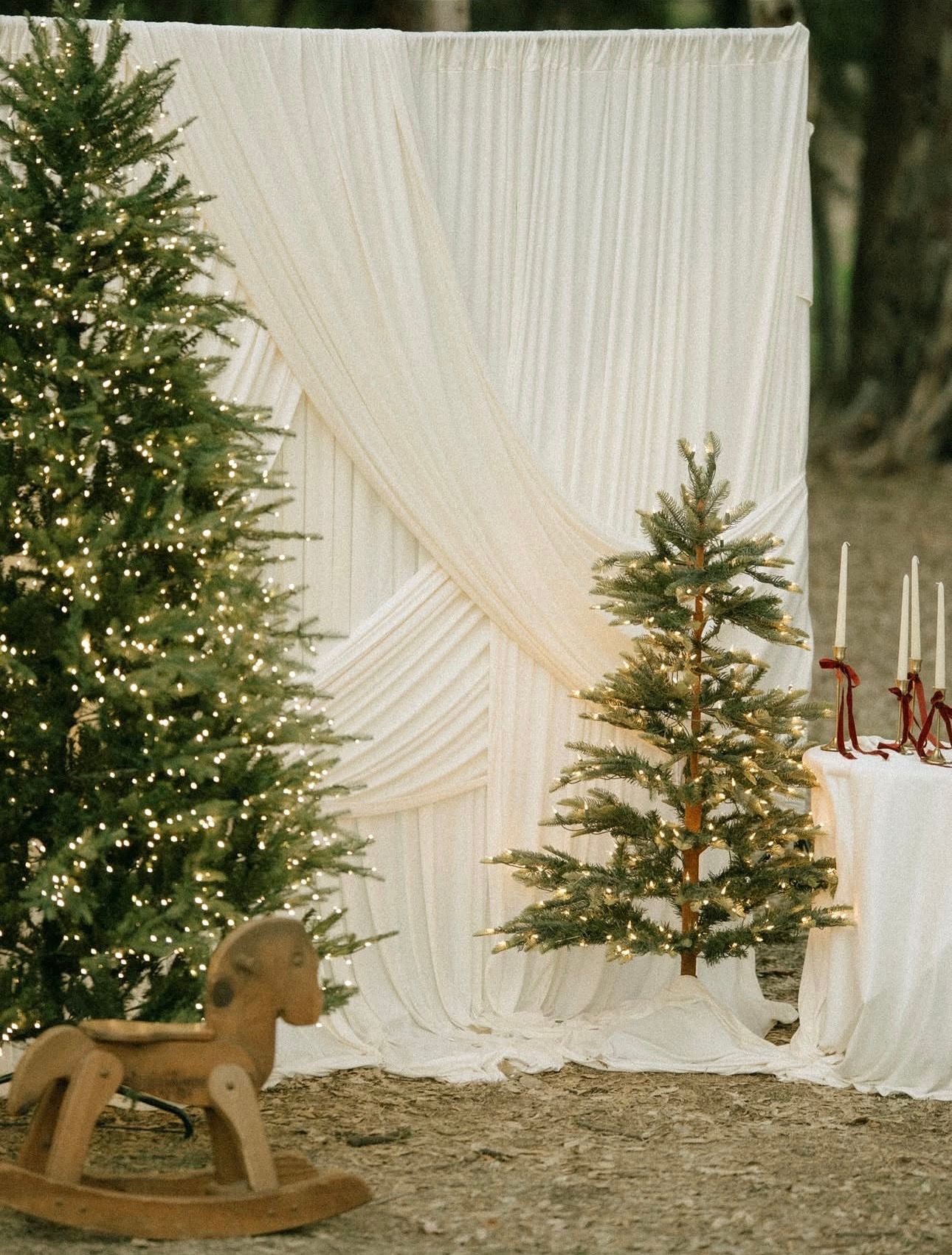 Elegant Christmas wedding backdrop with white draping, lit pine trees, wooden rocking horse, and candles on a decorated table.