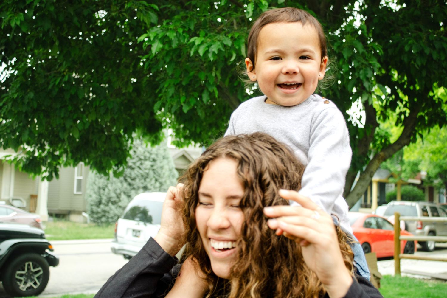 Smiling toddler sitting on a woman's shoulders under a leafy green tree outdoors.