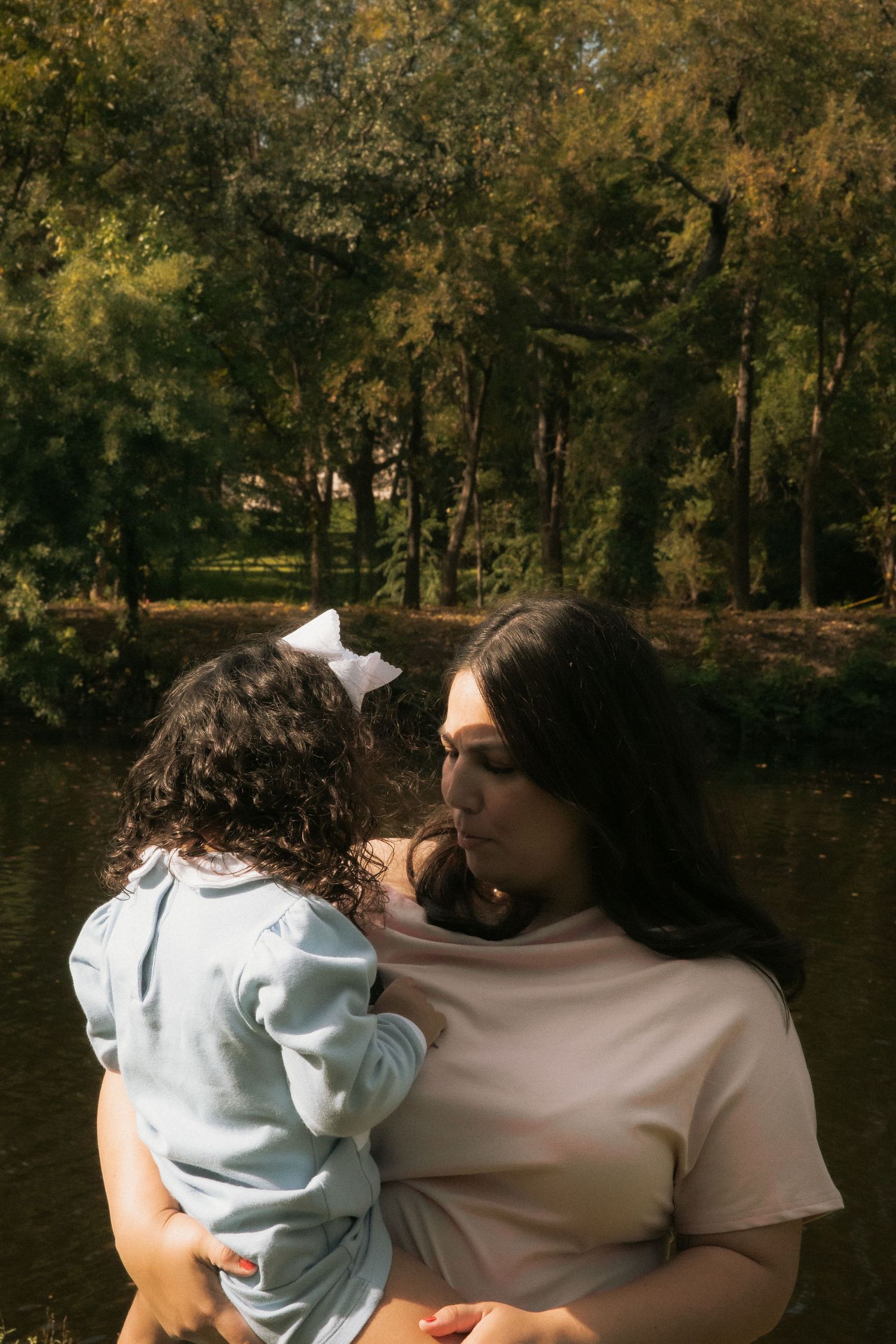 Mother tenderly holding her young daughter with a white bow near a sunlit pond surrounded by trees.