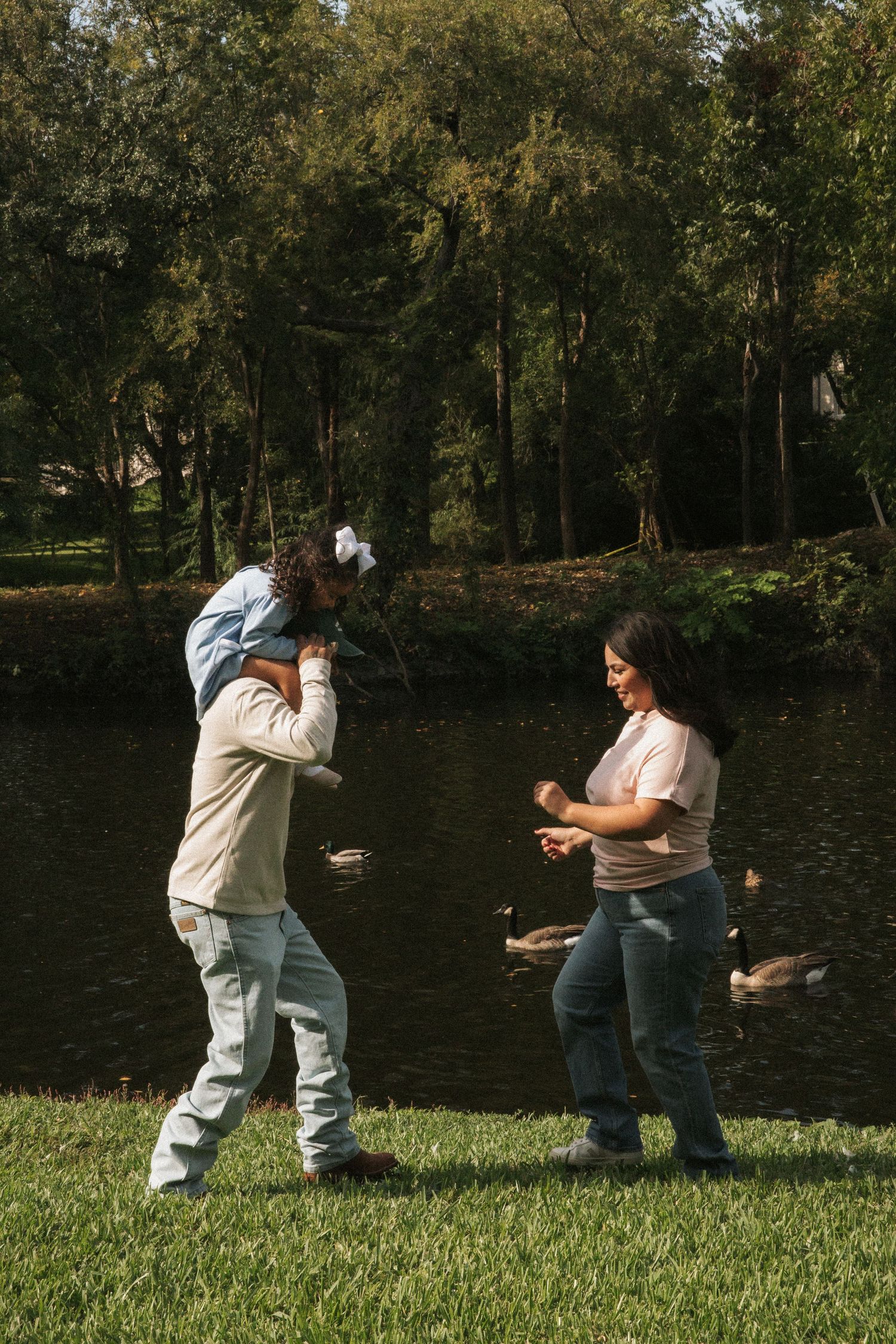 Family playing outdoors near a pond with ducks, father carrying child on shoulders.