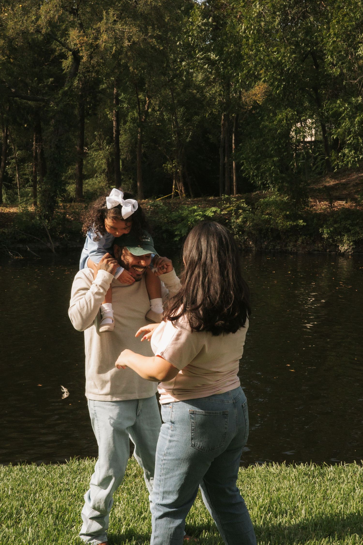 Family enjoying a sunny day by a pond in a lush green park, with a child on dad's shoulders.