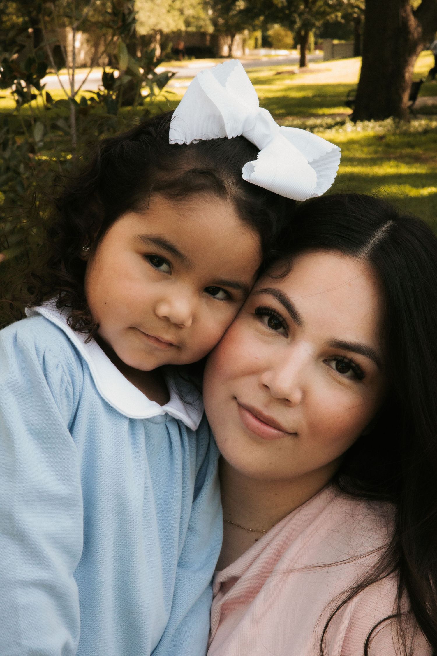 A mother and young daughter with a white bow share a tender cheek-to-cheek moment outdoors.