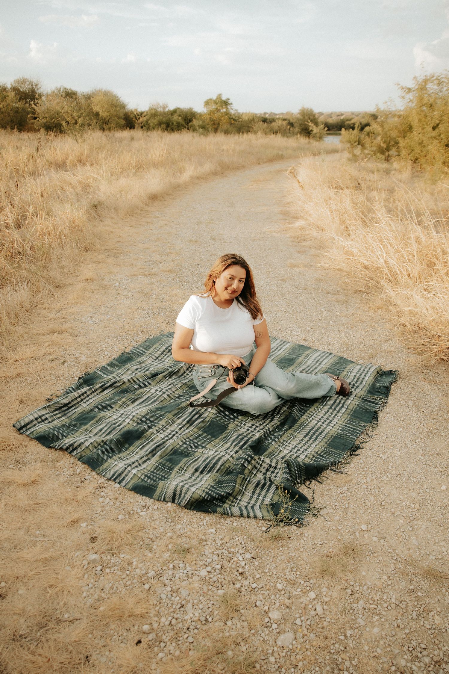 Woman sitting on plaid blanket on dirt path in golden field, holding camera, wearing white top and light jeans.