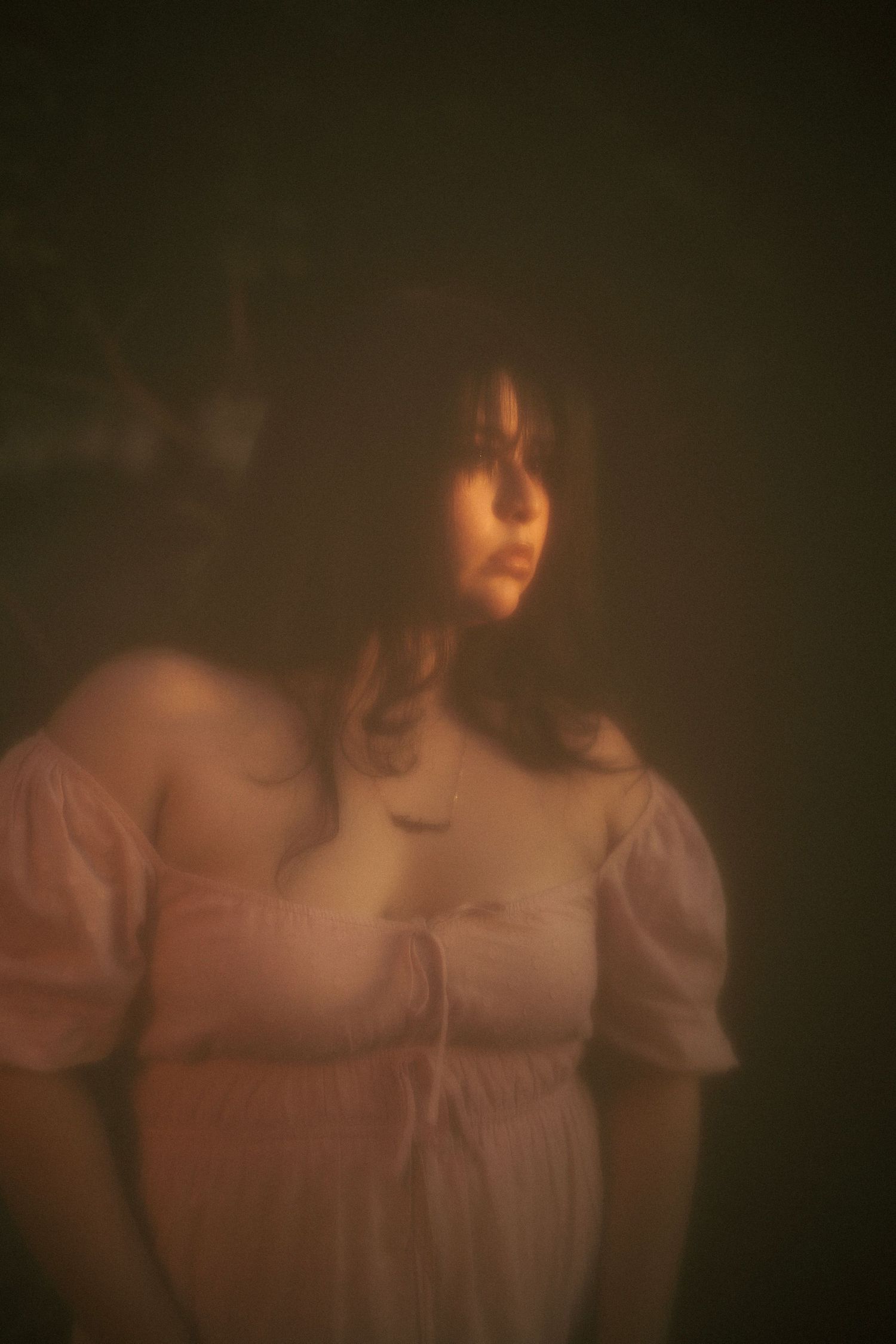 A young woman in a white dress stands in moody, warm-toned dim light with a dreamy, ethereal atmosphere.