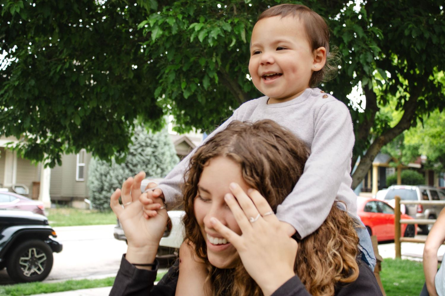Happy toddler boy riding on young woman's shoulders laughing outdoors under leafy trees.