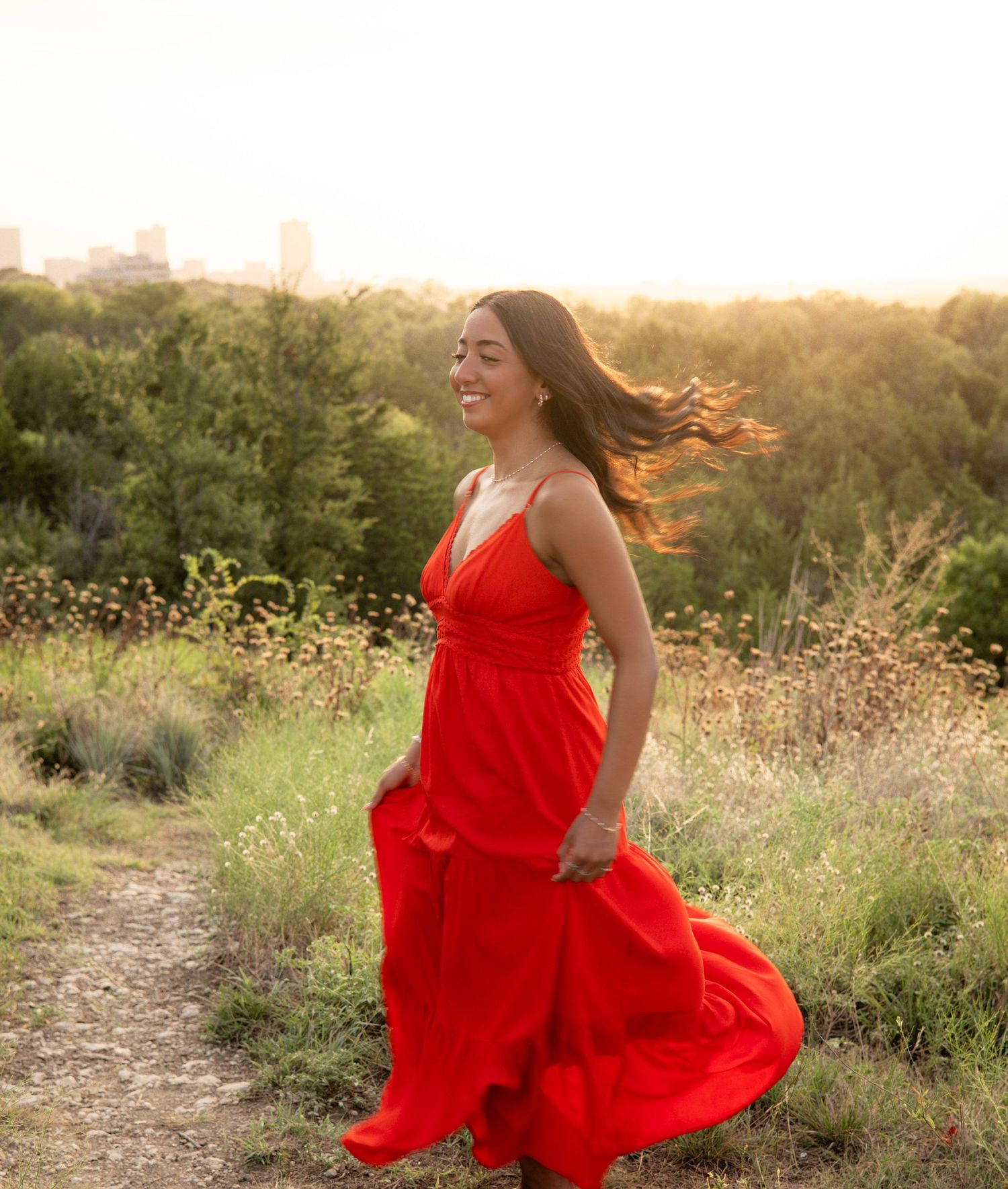 Young woman in red maxi dress spinning on a sunlit trail with green trees and city skyline.