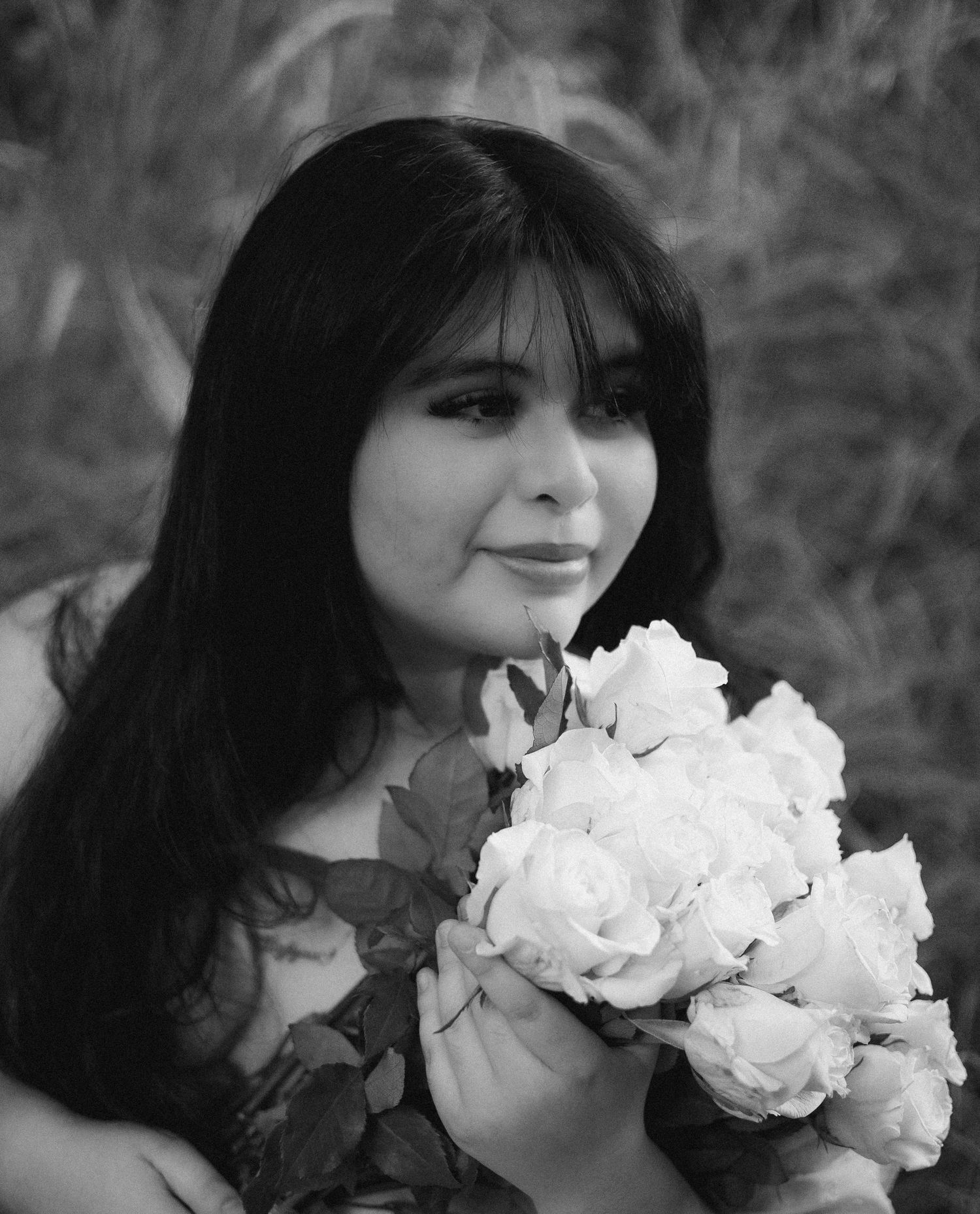 Black and white close-up portrait of young woman holding white roses bouquet outdoors.
