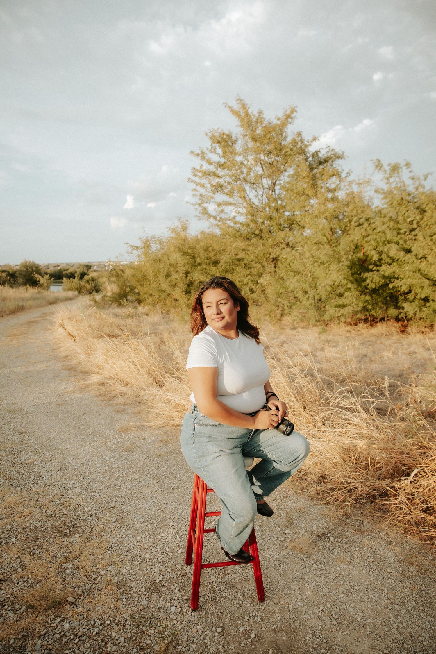 Woman in white tee and jeans sitting on red stool on a dirt path surrounded by dry grass and green trees.