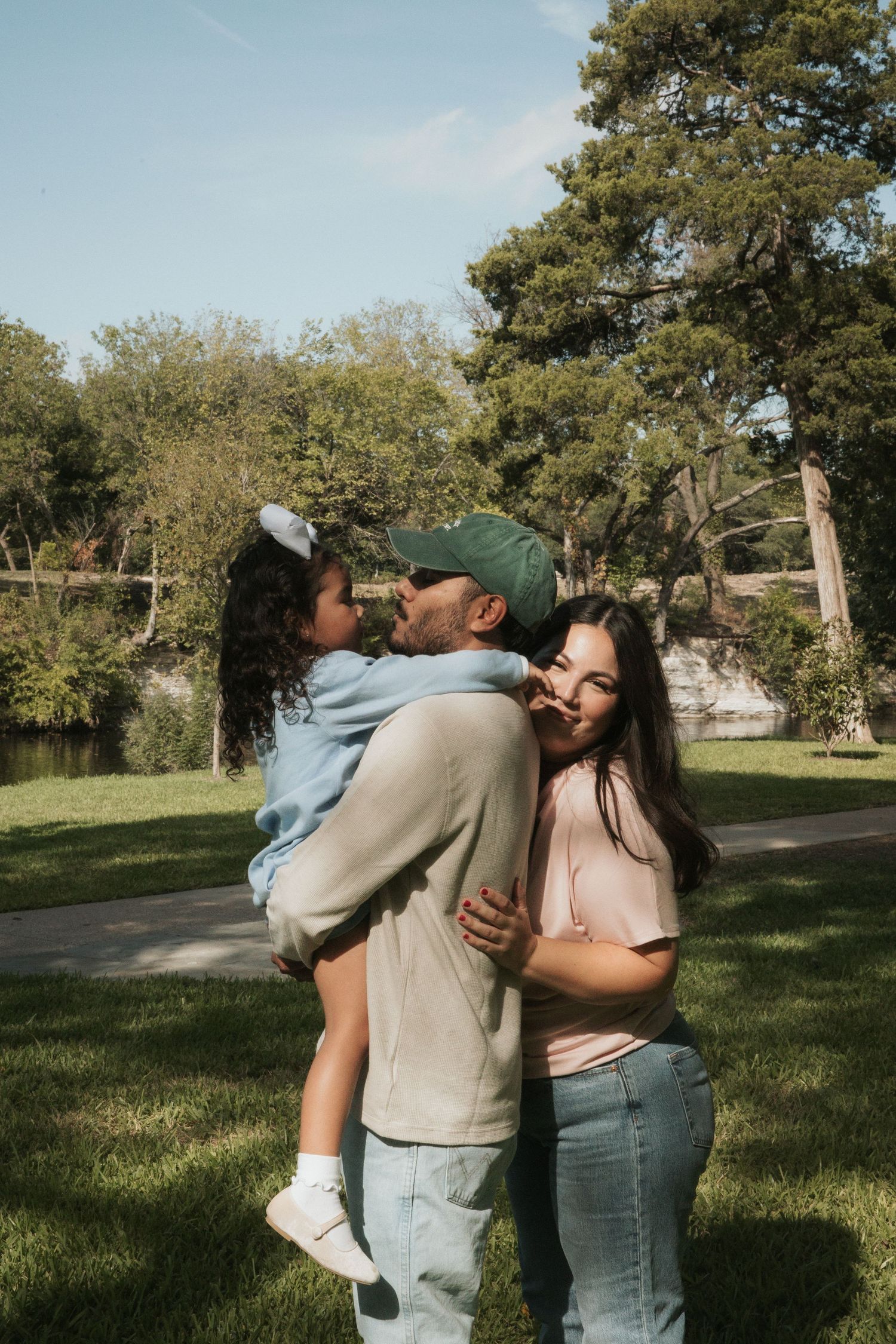 Happy family with father in green cap kissing daughter while mother embraces them in sunny park.