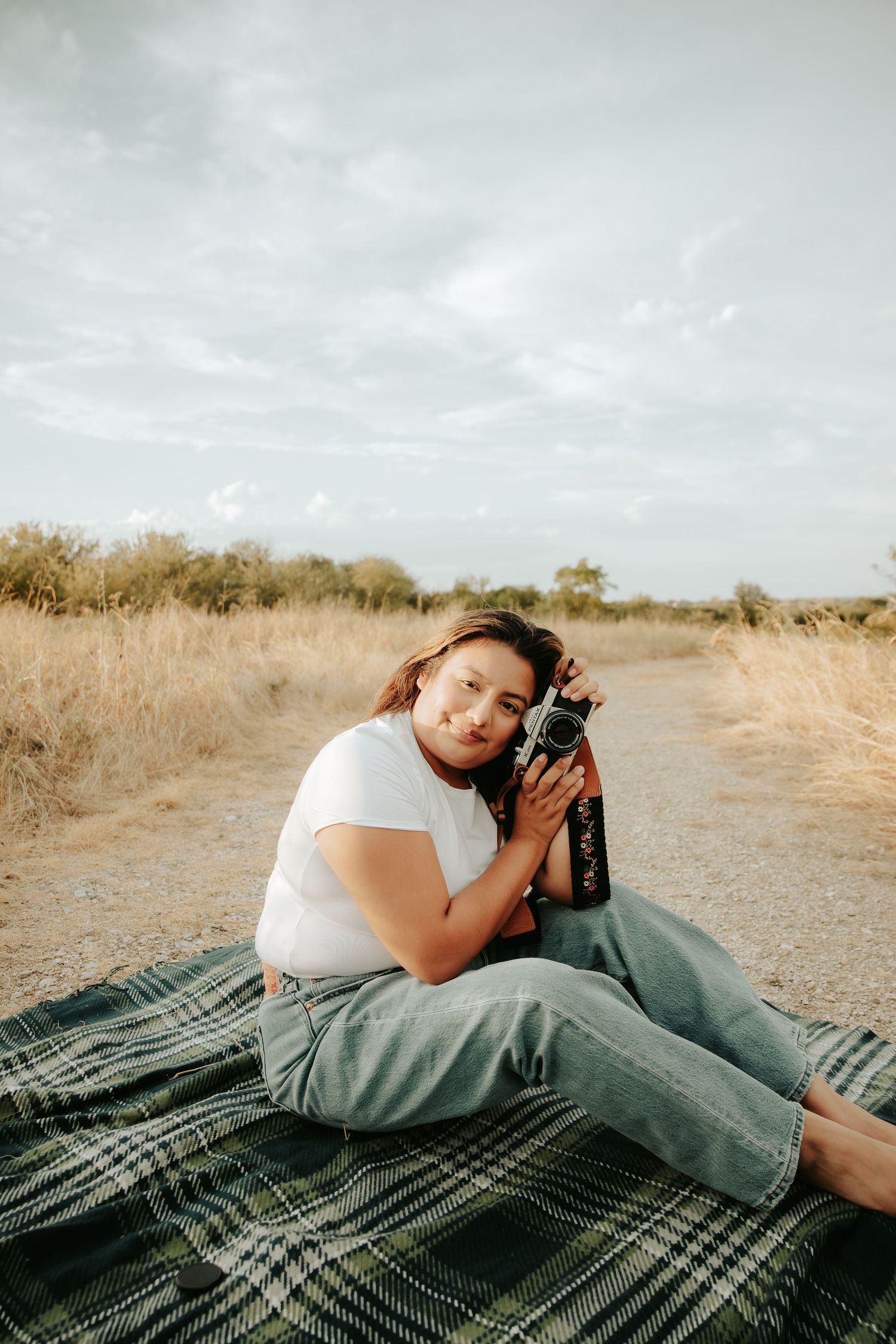 Young woman sitting on plaid blanket outdoors holding vintage camera in a dry field landscape.