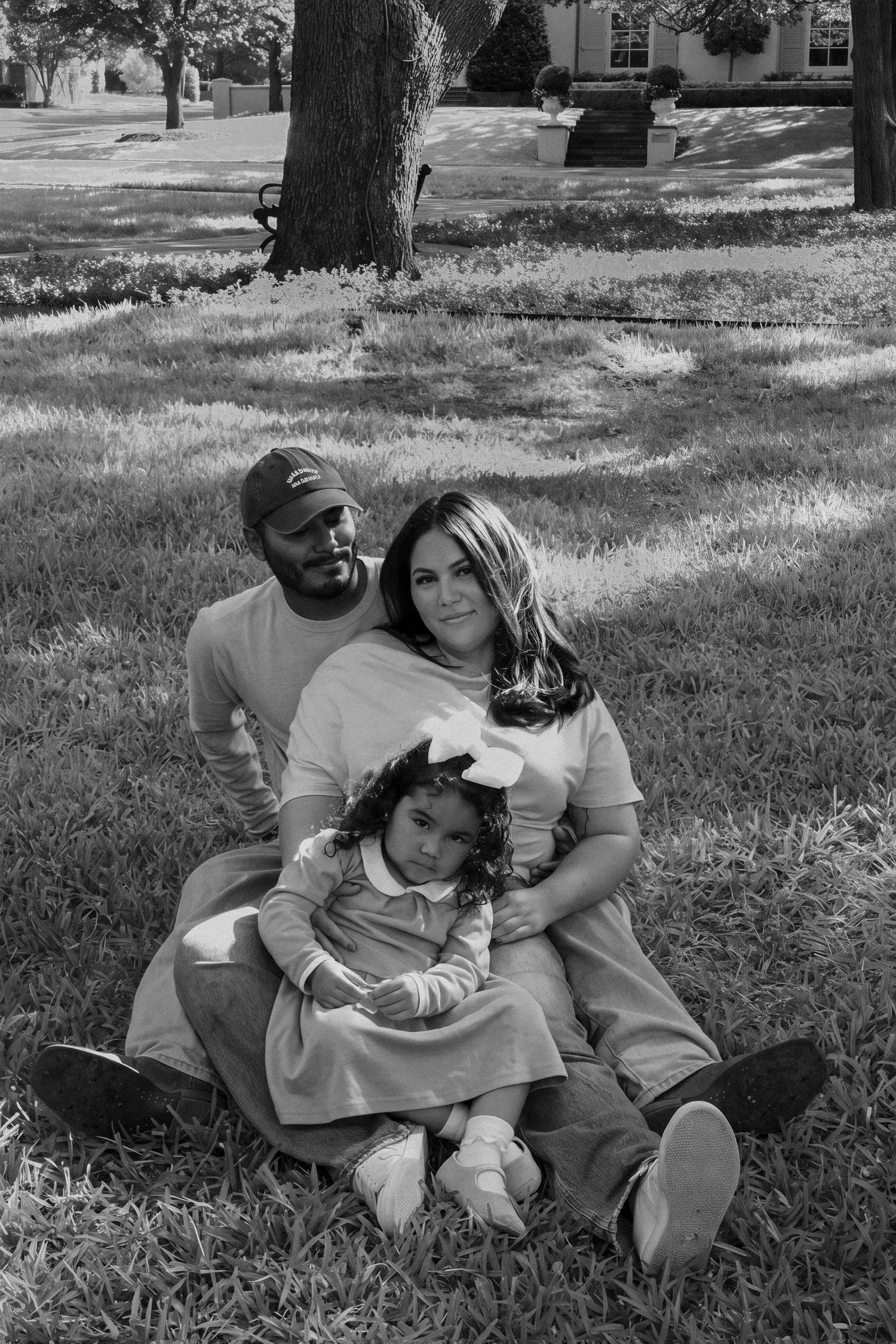 Black and white family portrait of parents and toddler sitting on grass in a park.