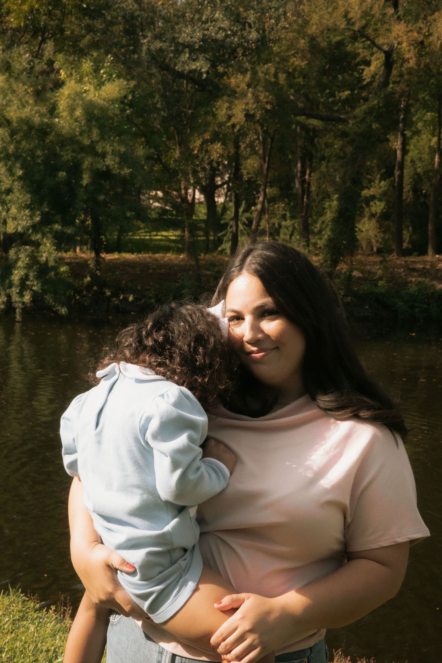 Smiling mother holding young daughter close while standing near a peaceful pond in an autumn park.