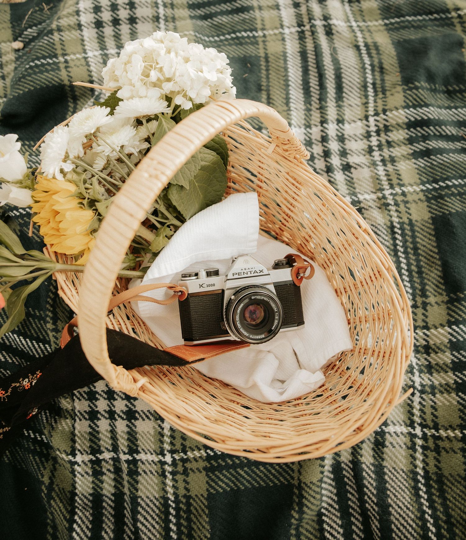 A vintage film camera resting in a wicker basket with white flowers on a plaid blanket.
