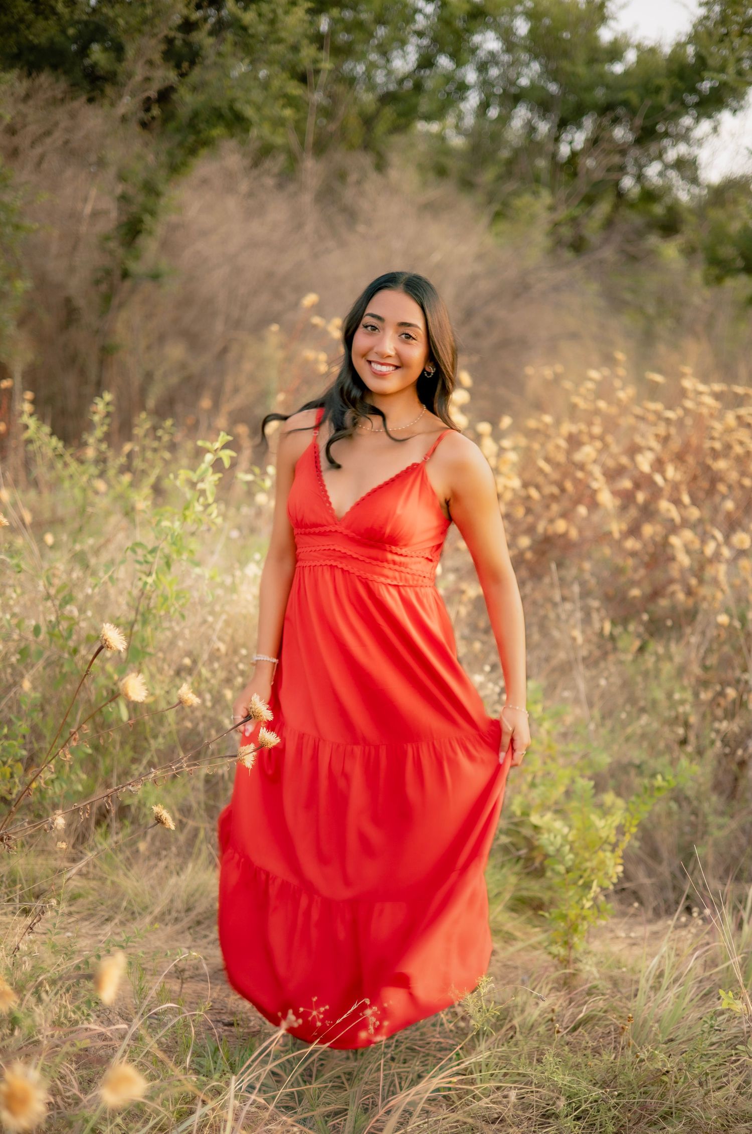 Young woman in red maxi dress standing and smiling in a golden wildflower field.