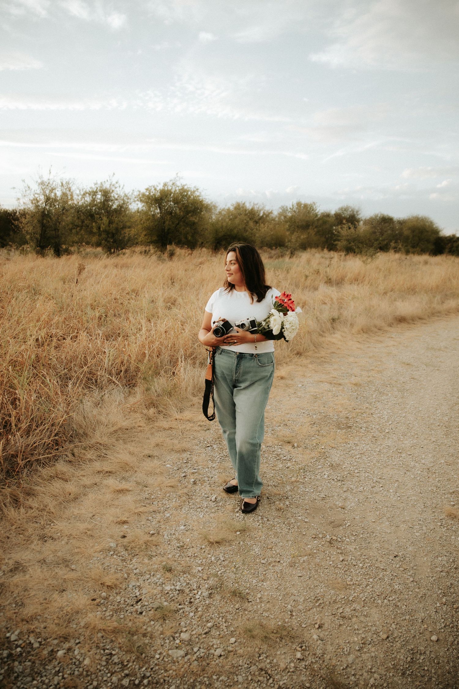 Woman walking on dirt path holding flowers and camera, surrounded by golden dry grass fields under cloudy sky.