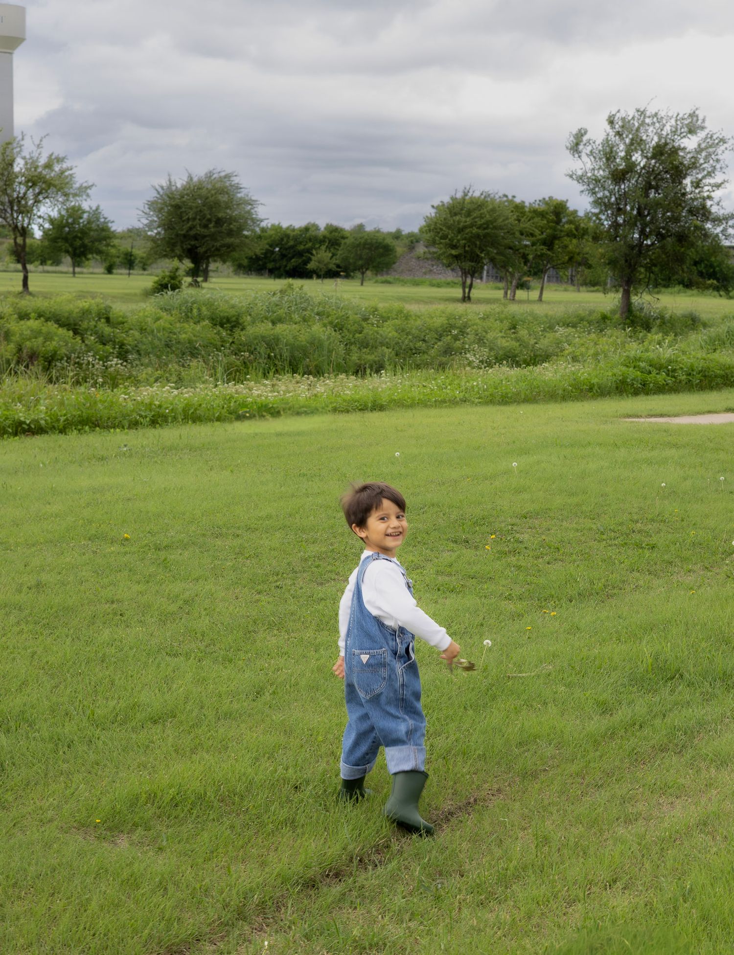 Happy toddler boy in denim overalls and green rain boots running on green lawn.