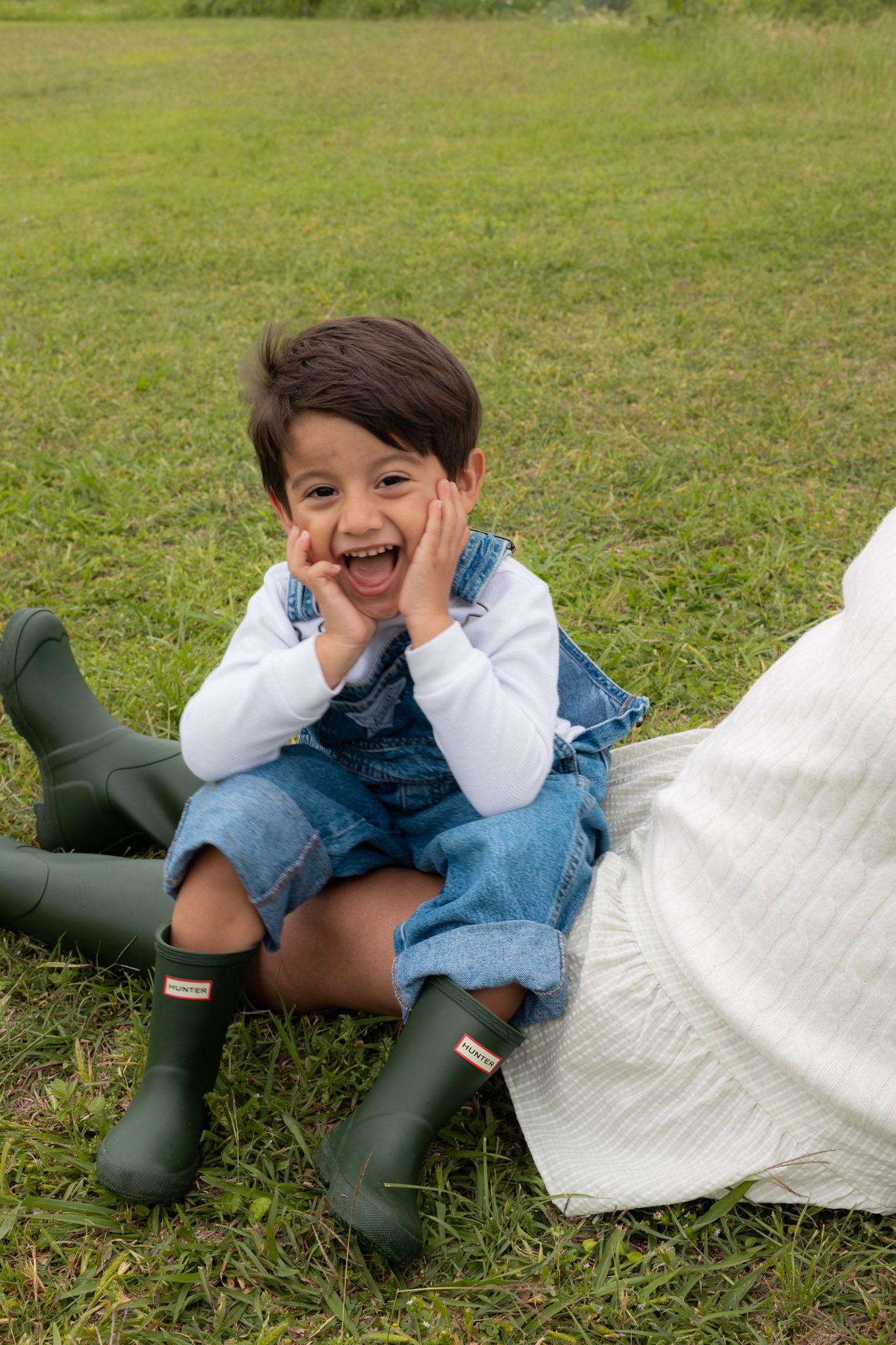 Laughing toddler in denim overalls and green Hunter boots sitting on green grass outdoors.