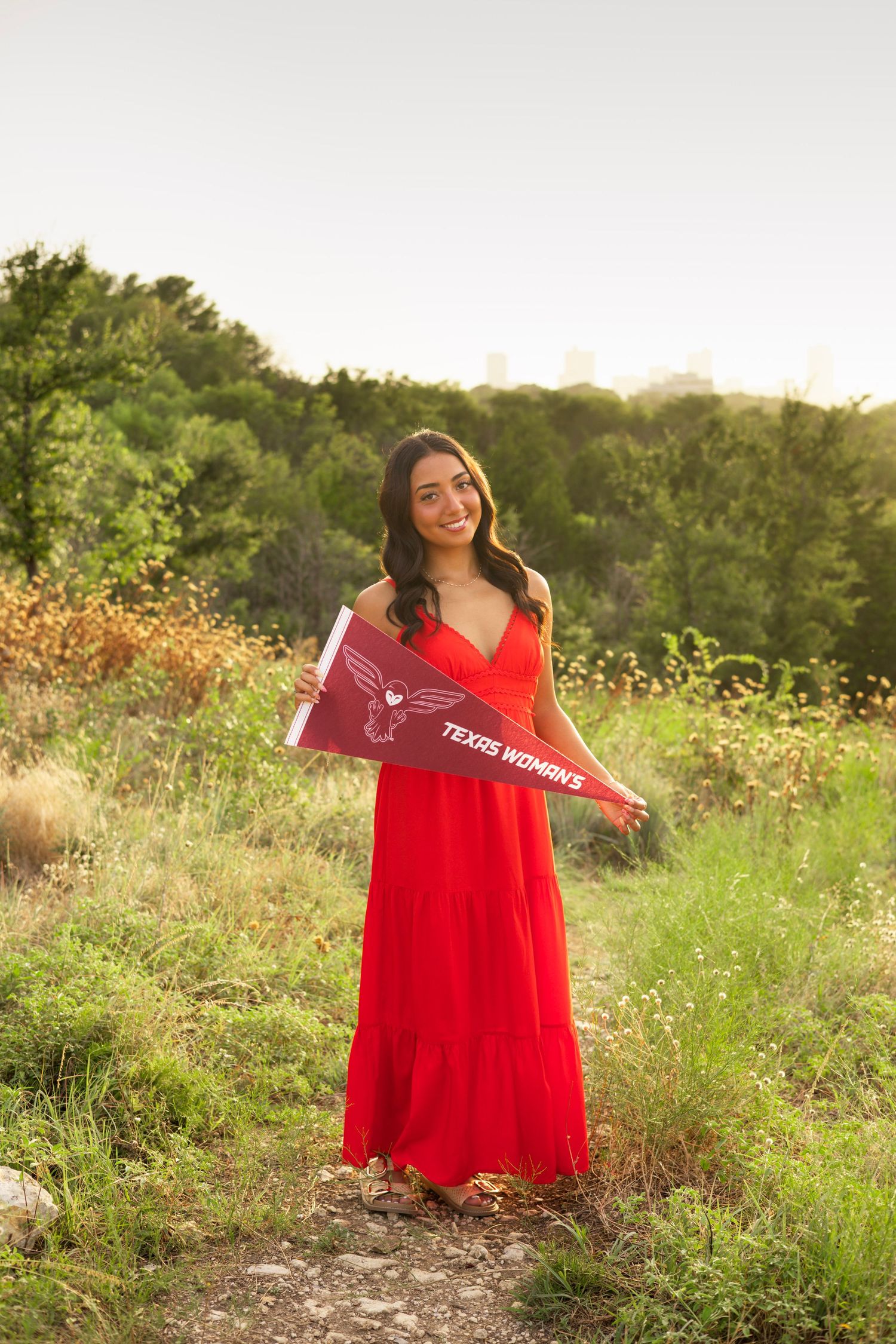 Young woman in red maxi dress holding Texas Woman's University pennant outdoors at golden hour.