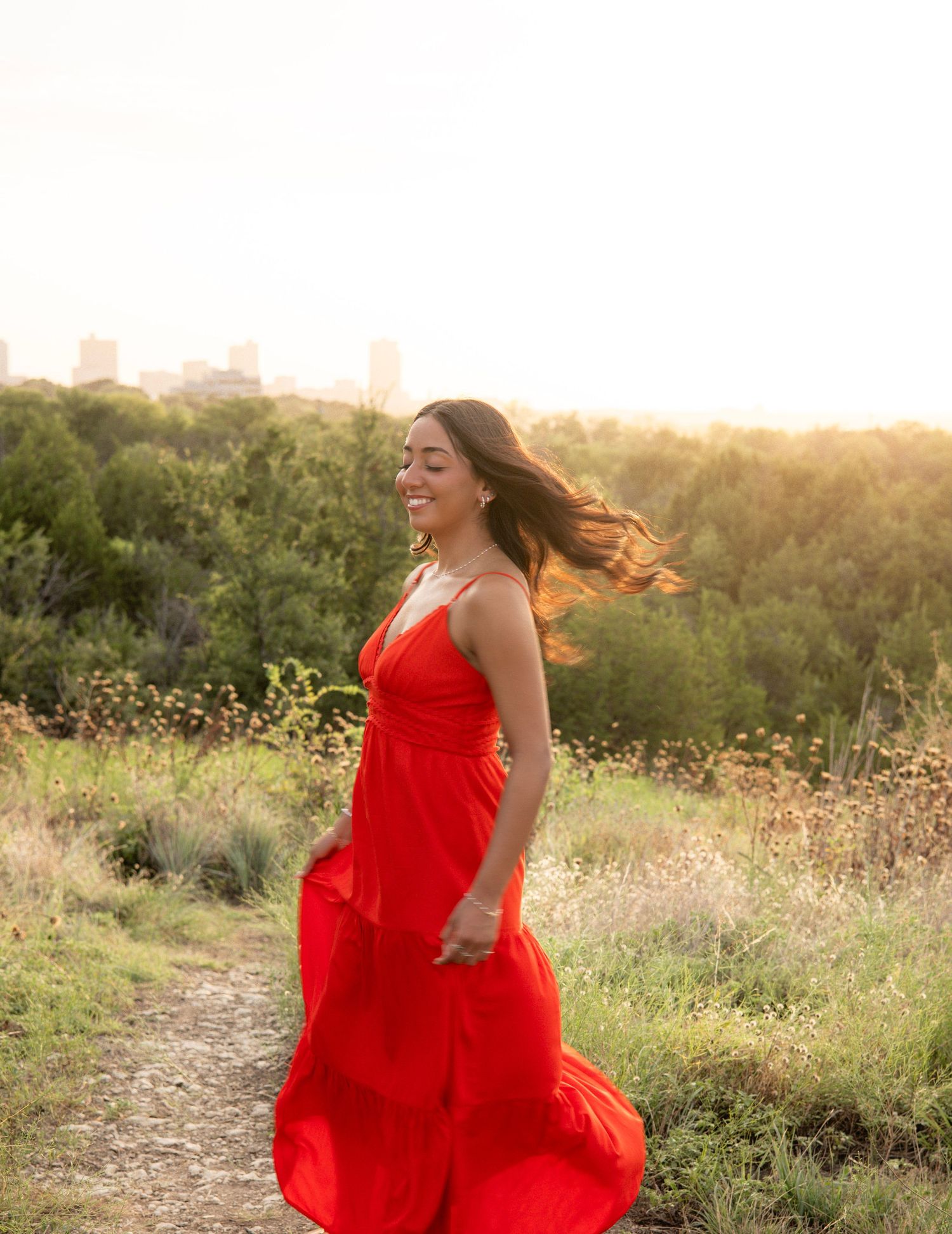 Joyful woman in flowing red dress spinning on a sunlit path with city skyline in background.
