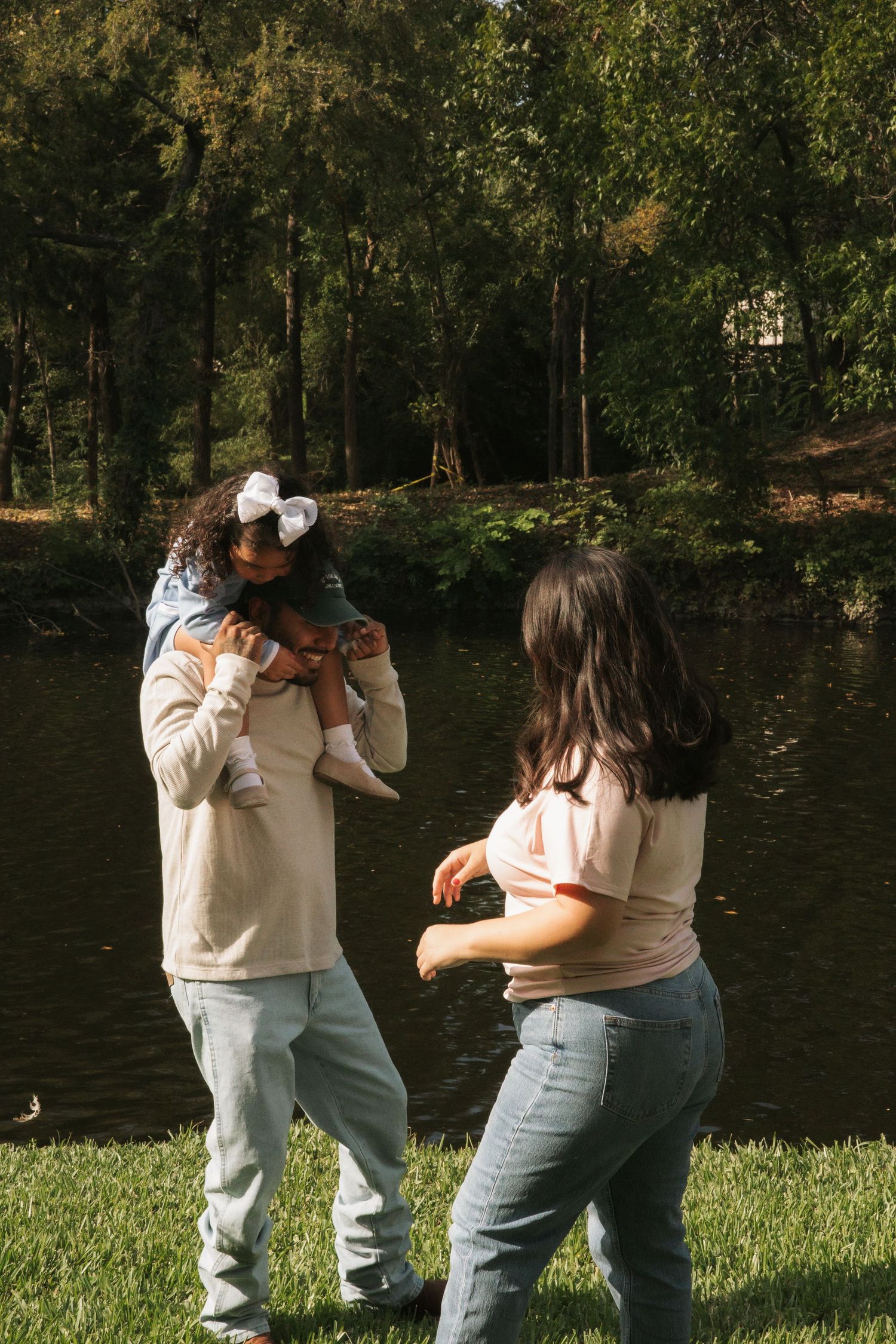Father holding young daughter with white bow near pond as mother reaches out playfully in sunny park.