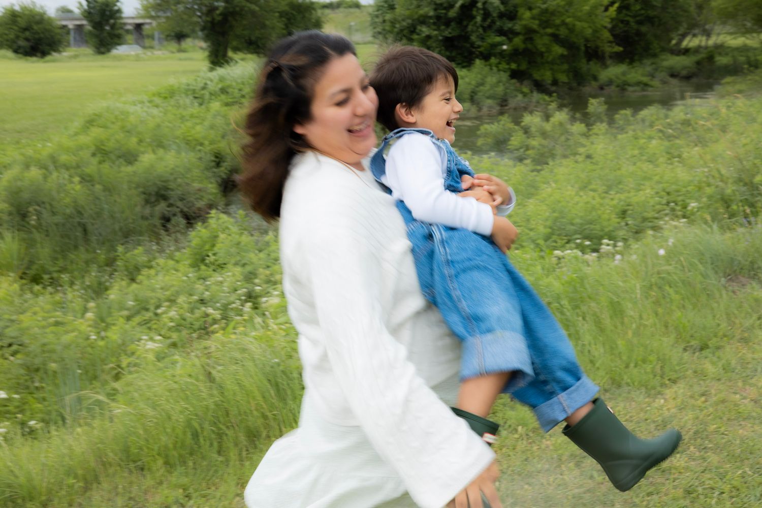 A smiling mother in a white coat carries a laughing toddler in denim overalls through a lush green field.