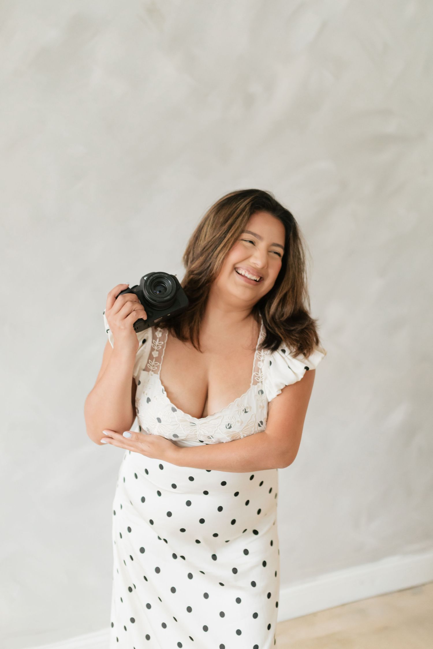 Smiling female photographer in white polka dot dress holding camera against gray wall.