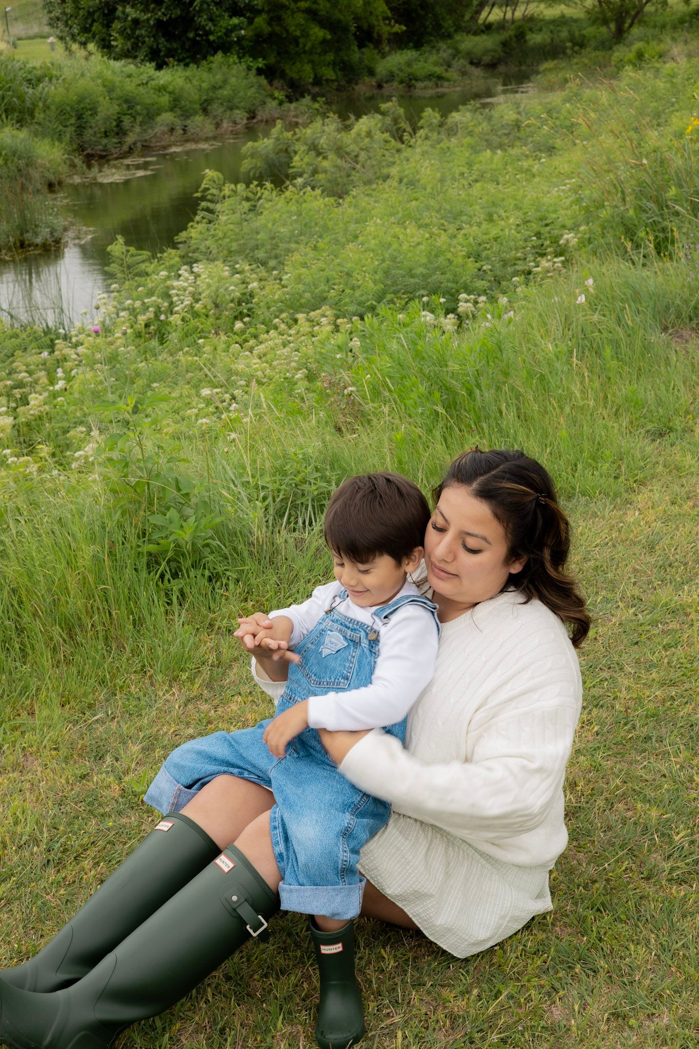 A mother in a white sweater and green boots sits on grass holding her toddler in denim overalls near a pond.