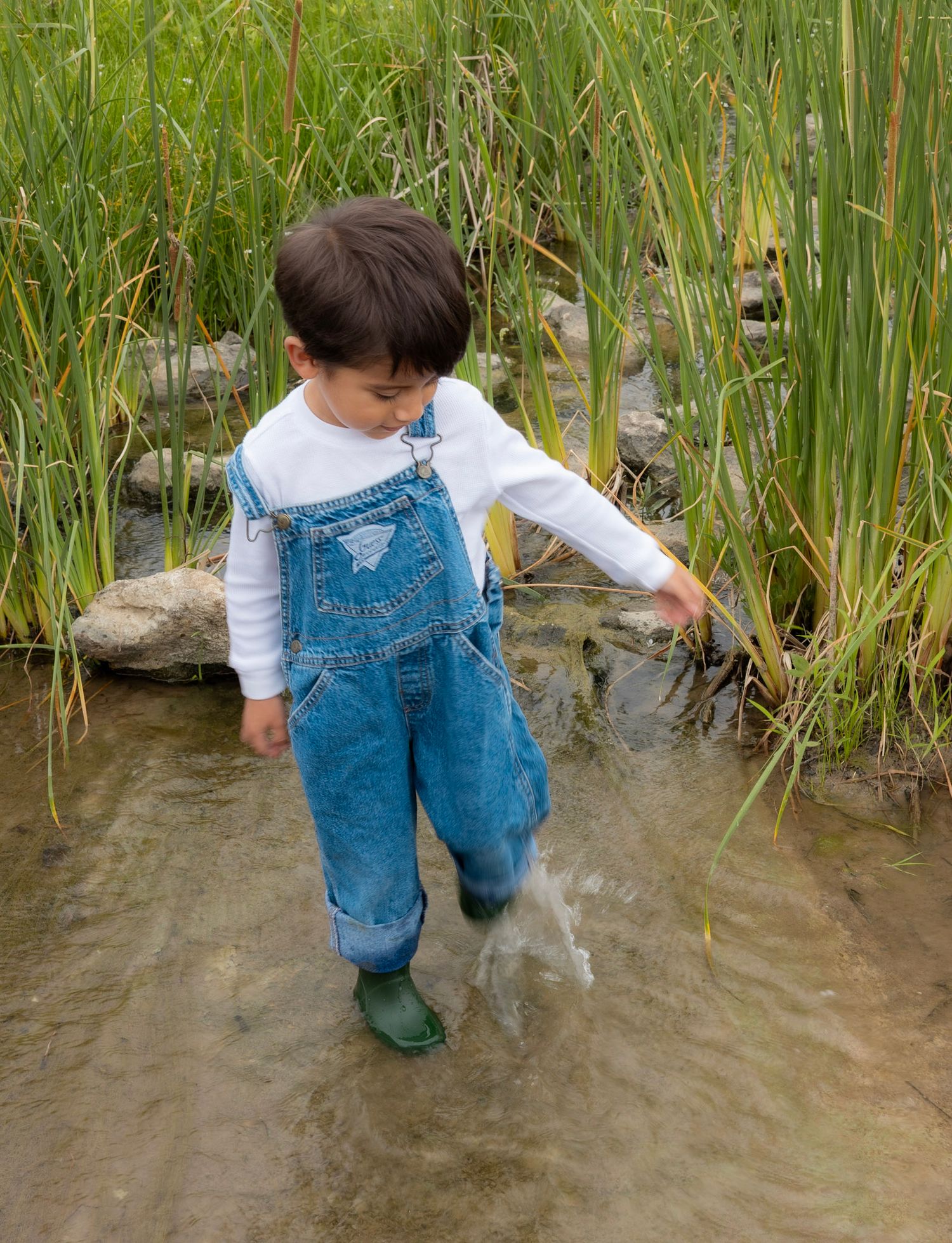 Young child in denim overalls and green boots wading through a shallow stream near tall grass.