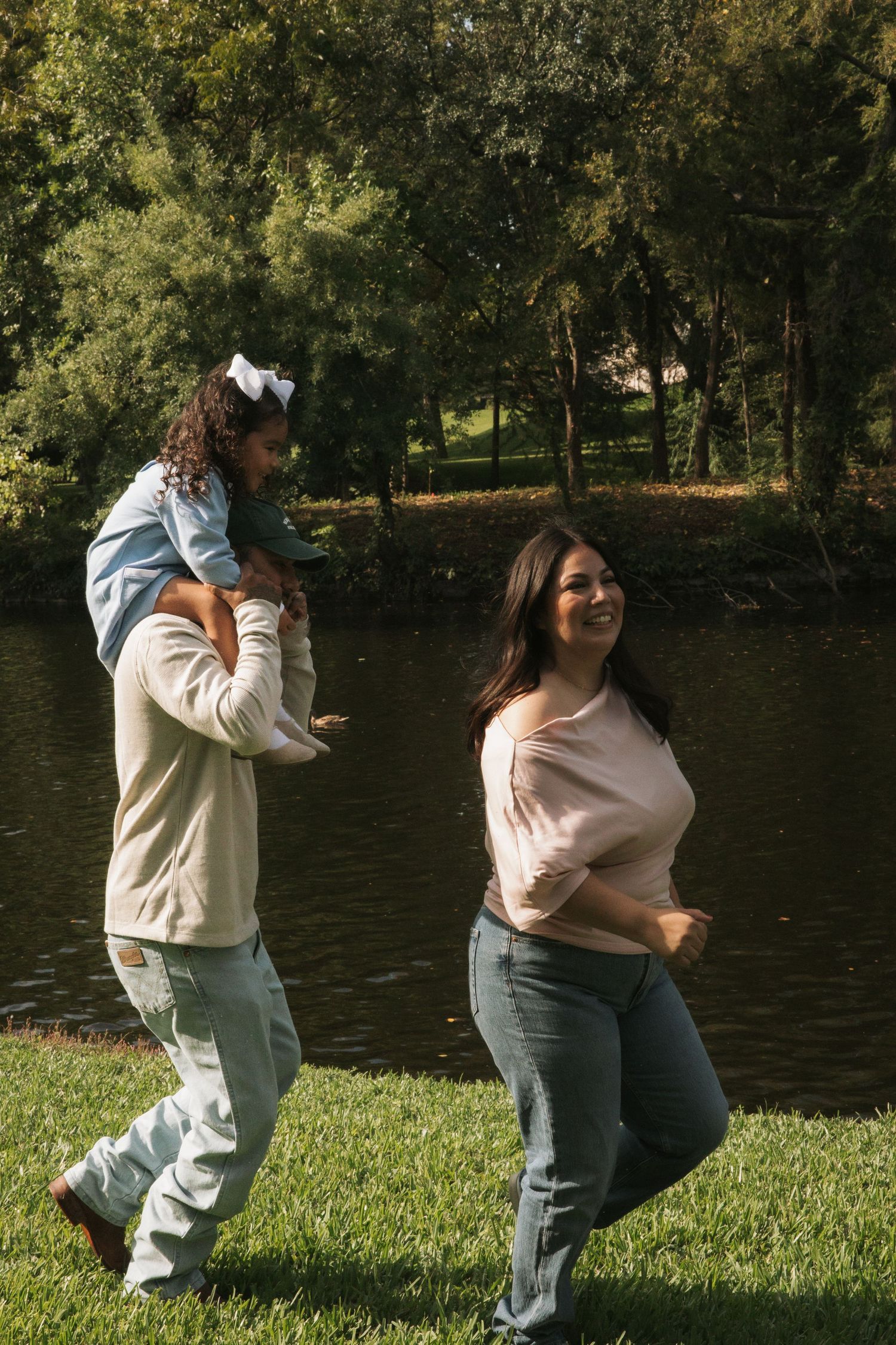 Family walking by a serene pond in a sunny park, man carrying child on shoulders with smiling woman beside them.