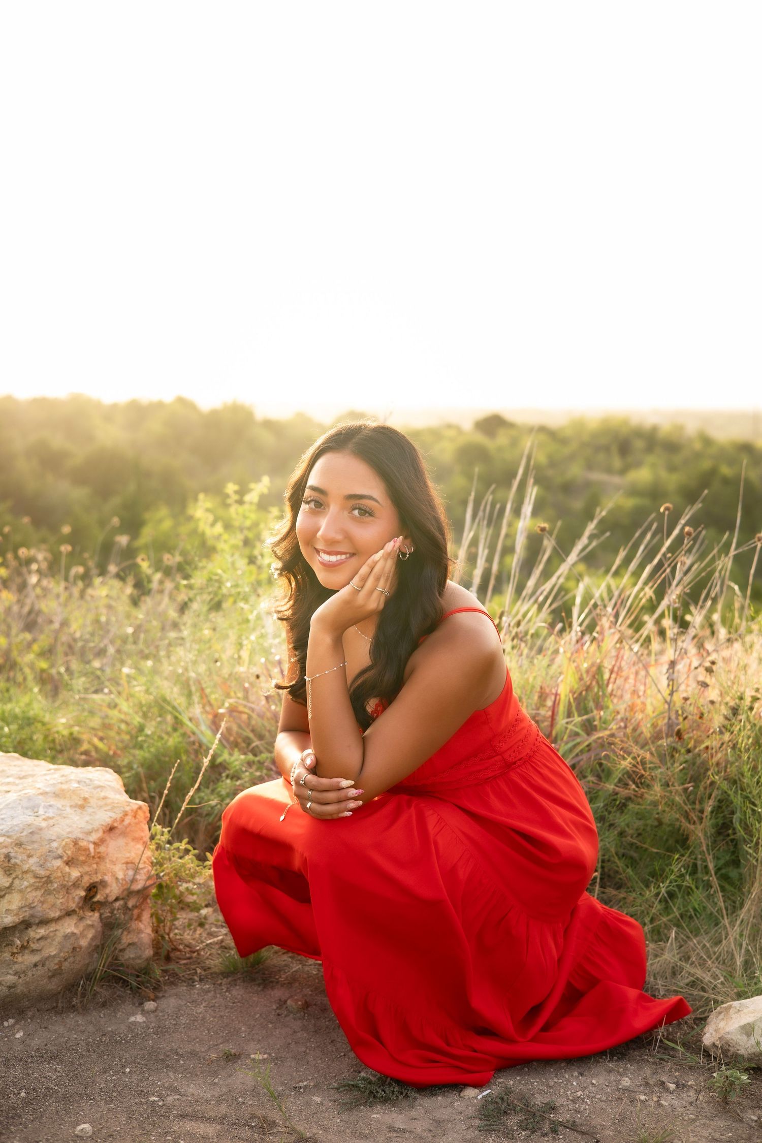 Smiling young woman in red dress sitting on rocks in golden hour sunlit field.