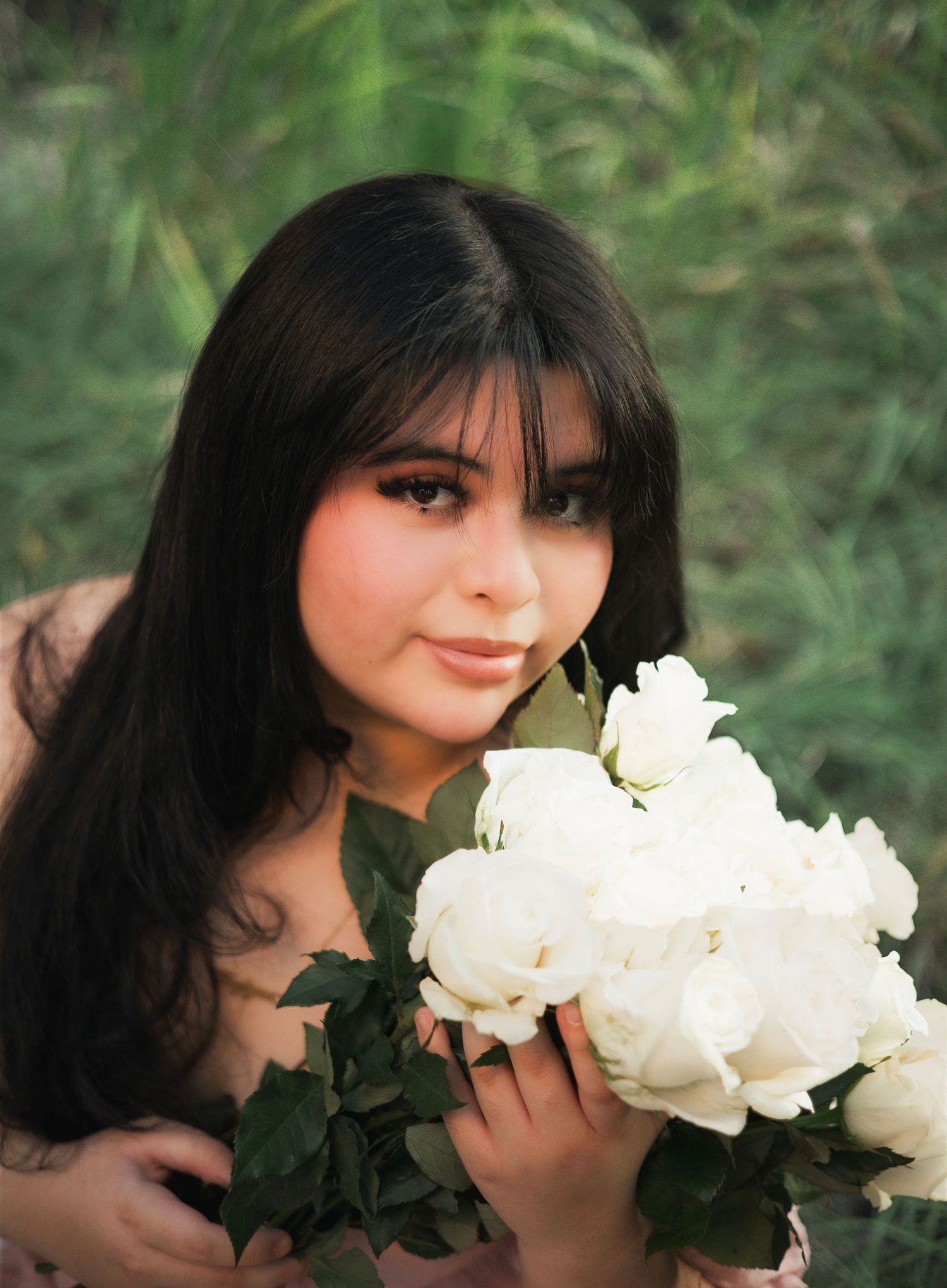 Young woman with long dark hair holding white roses bouquet close to face in lush greenery.