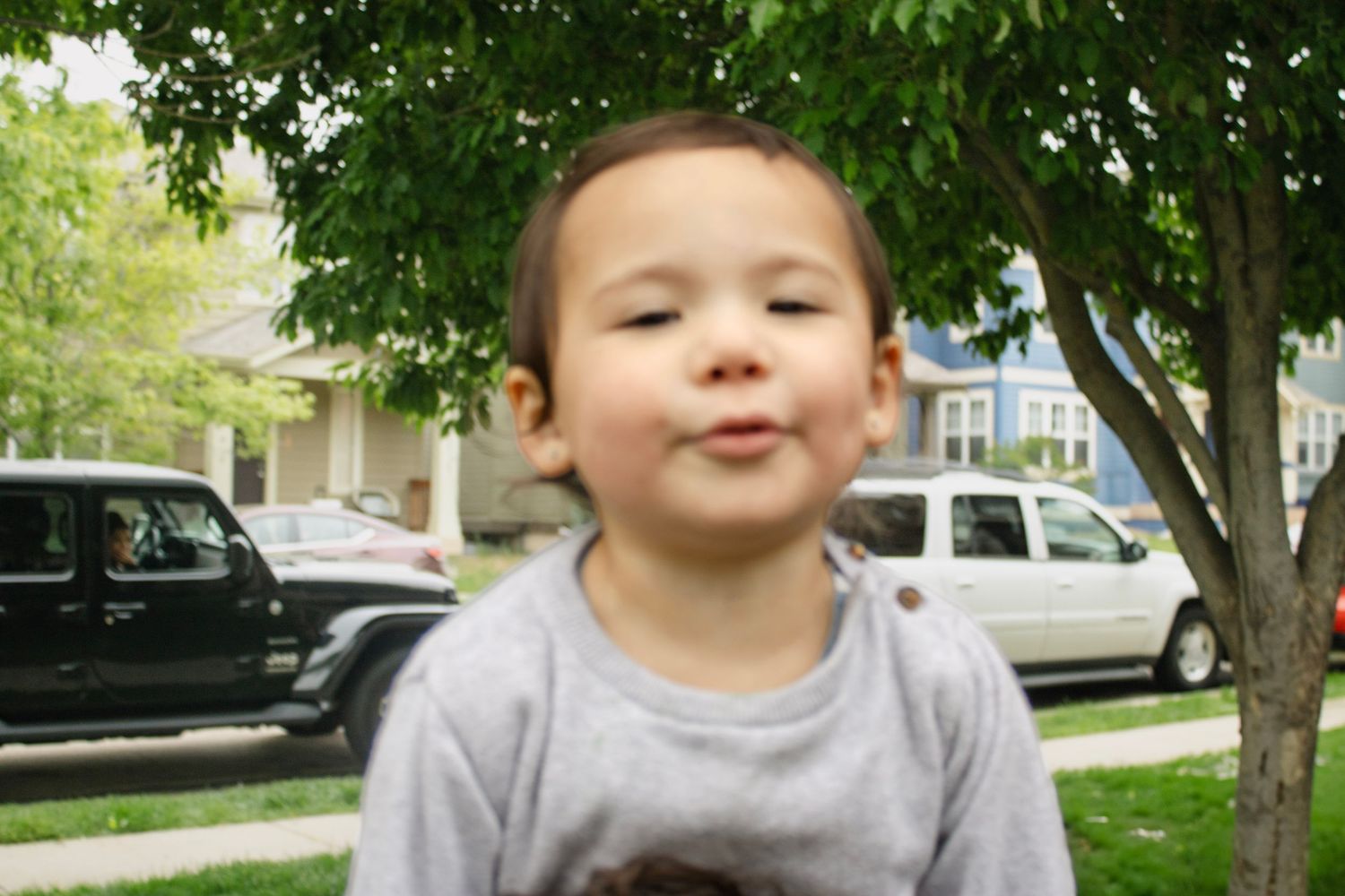 Cute toddler boy making funny face standing near tree in suburban neighborhood setting.