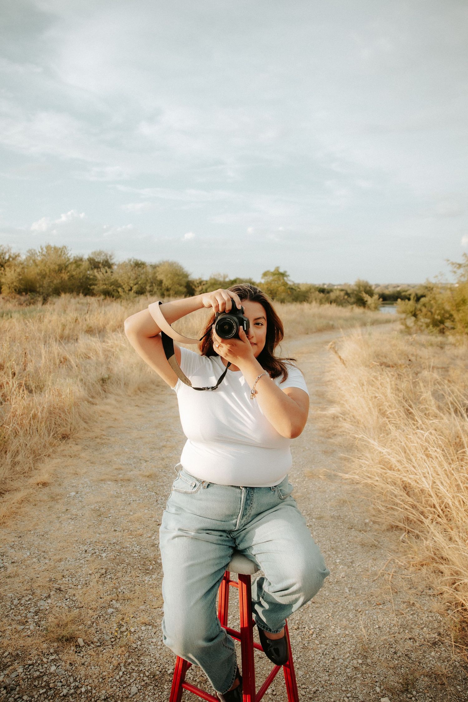 Woman in white top and jeans sits on red stool on dirt path, holding camera up to photograph in golden field.