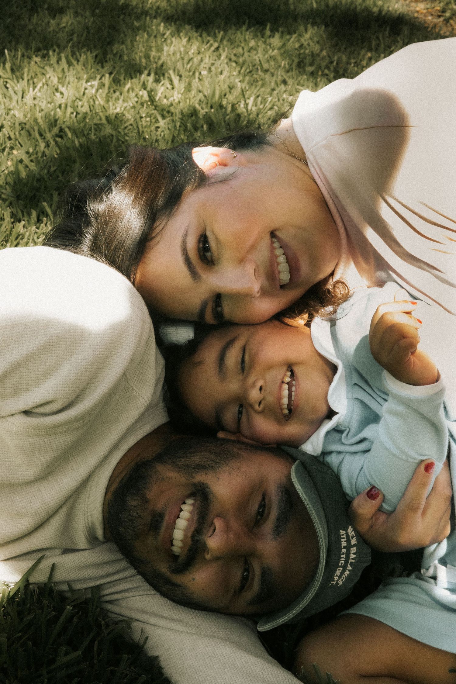 Happy family of three lying on green grass smiling up at camera on a bright sunny day.