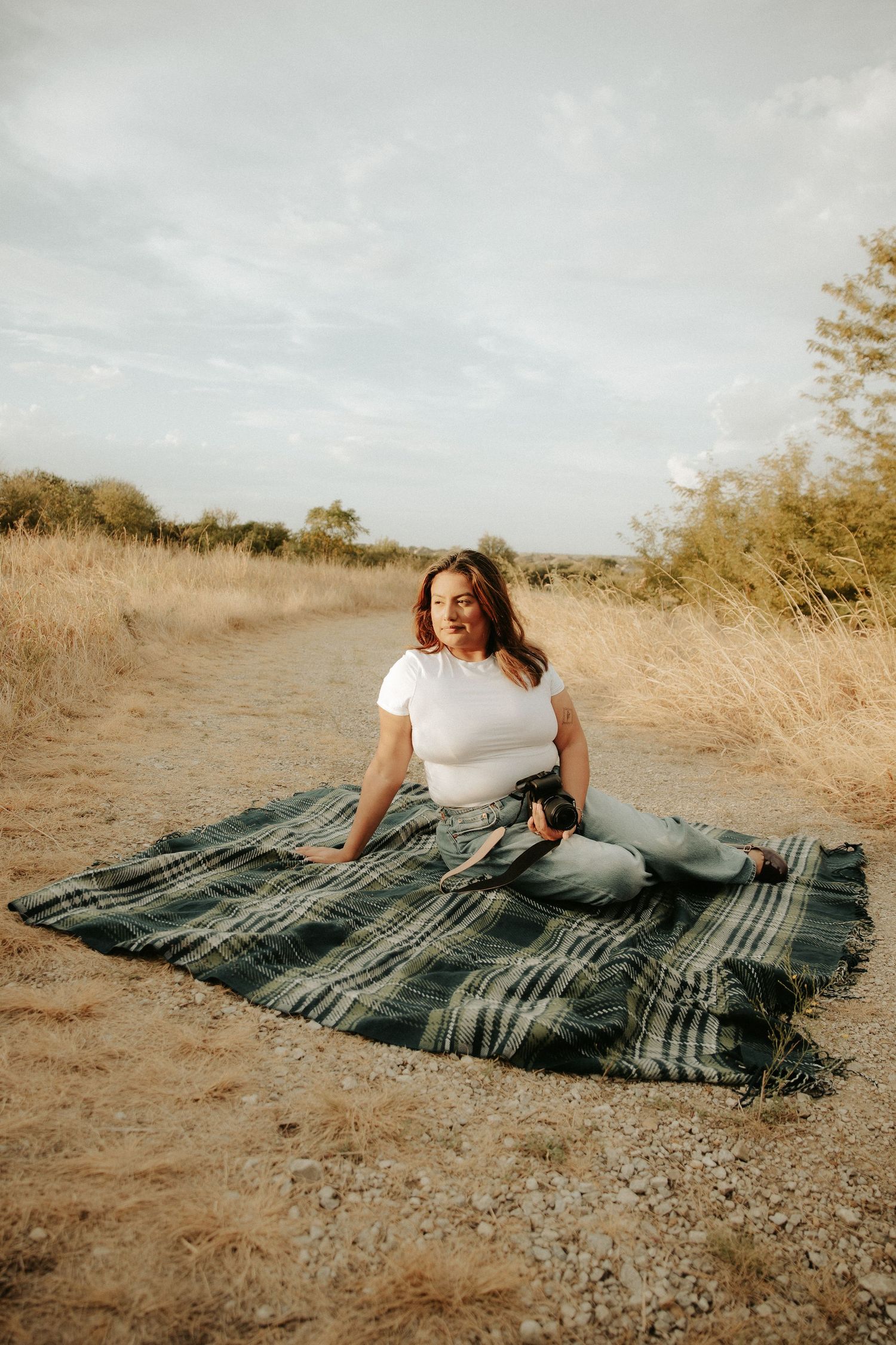 Young woman in white t-shirt and jeans relaxes on plaid blanket on rustic dirt path surrounded by dry grass.