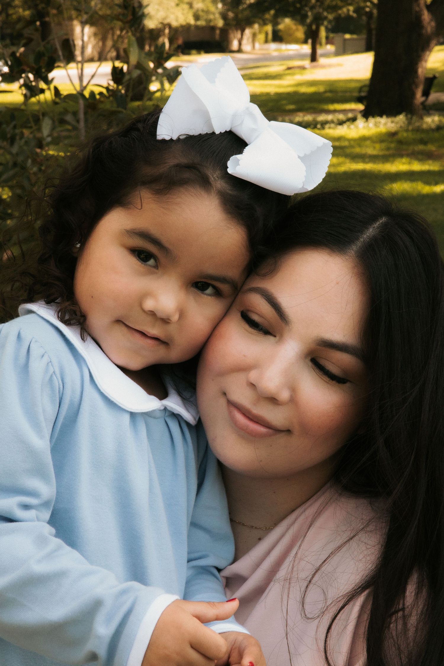 Tender moment between mother and young curly-haired daughter with white bow in an outdoor park setting.