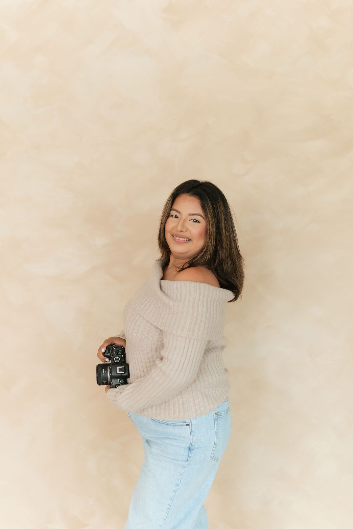 Smiling woman holding a camera, wearing an off-shoulder beige sweater and light jeans, posed against a warm neutral backdrop.