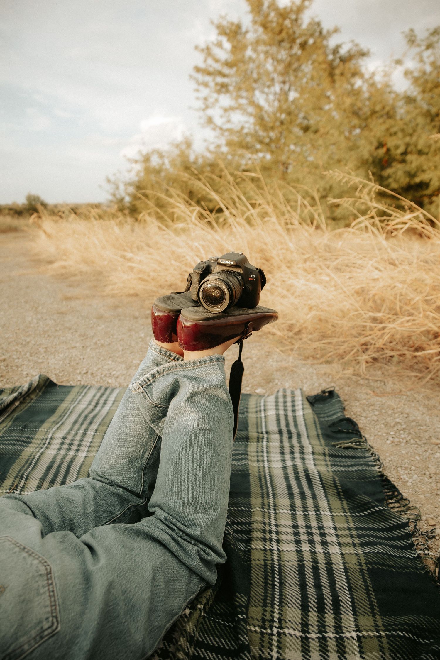 Person relaxing on plaid blanket outdoors with camera balanced on red boots in dry grassy field.