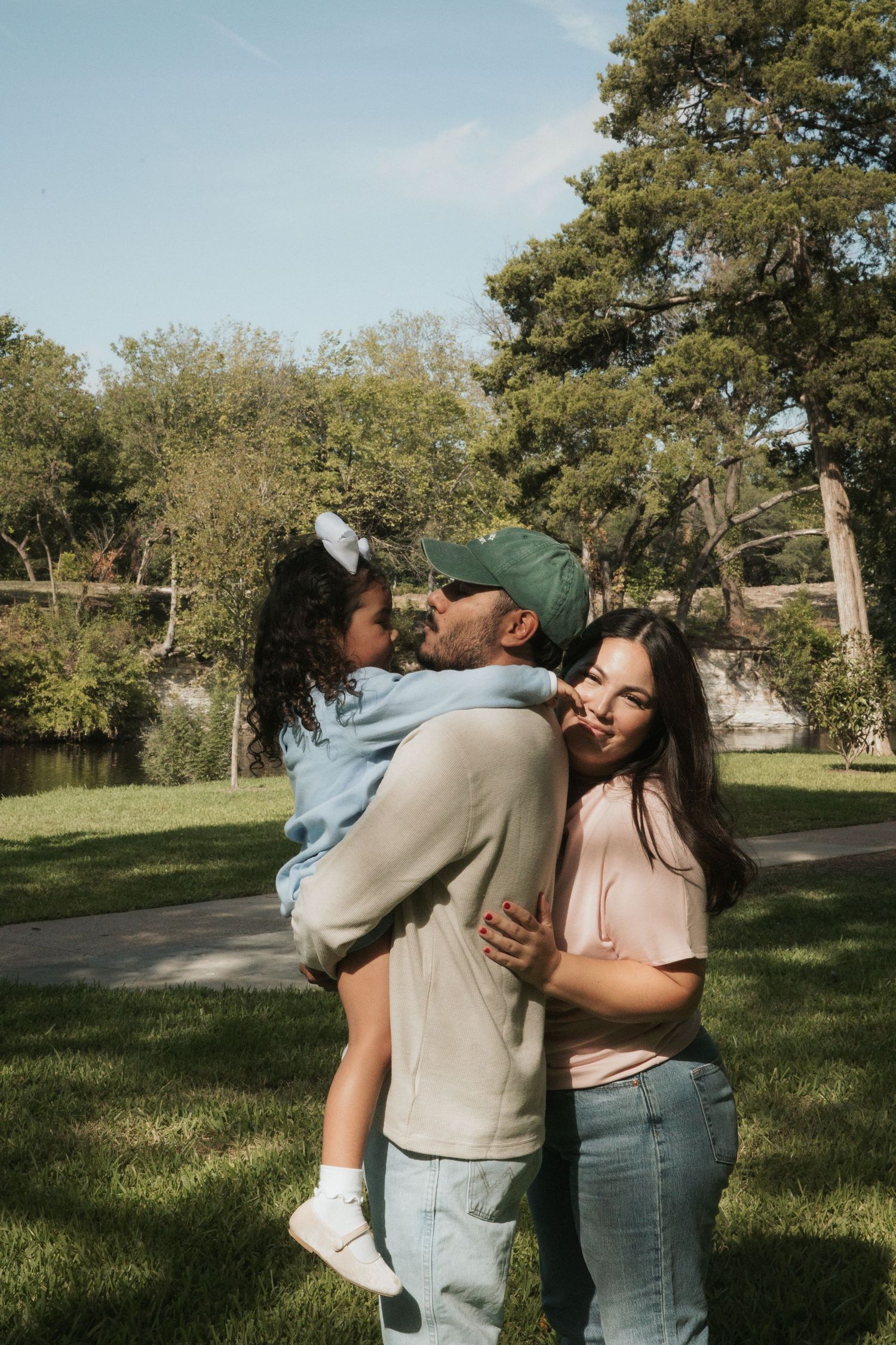 A family of three shares a tender moment in a park, with a father kissing his young daughter while mother smiles nearby.