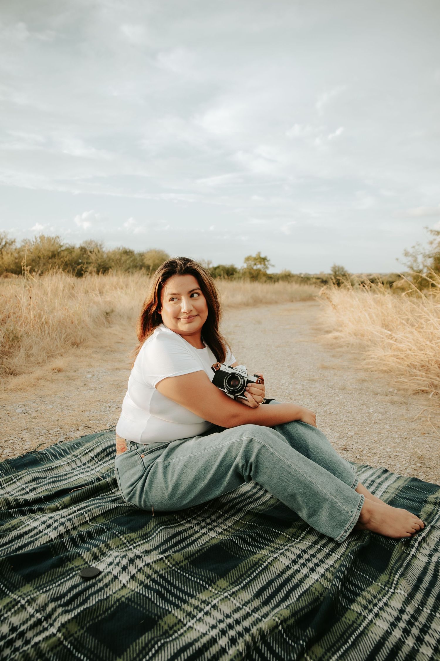 Smiling woman in white tee and jeans sits barefoot on plaid blanket outdoors holding a camera.