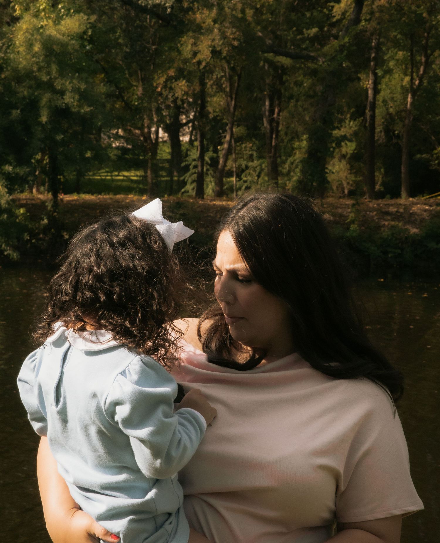 Mother gazing lovingly at young curly-haired daughter with white bow near a sunlit pond in park.