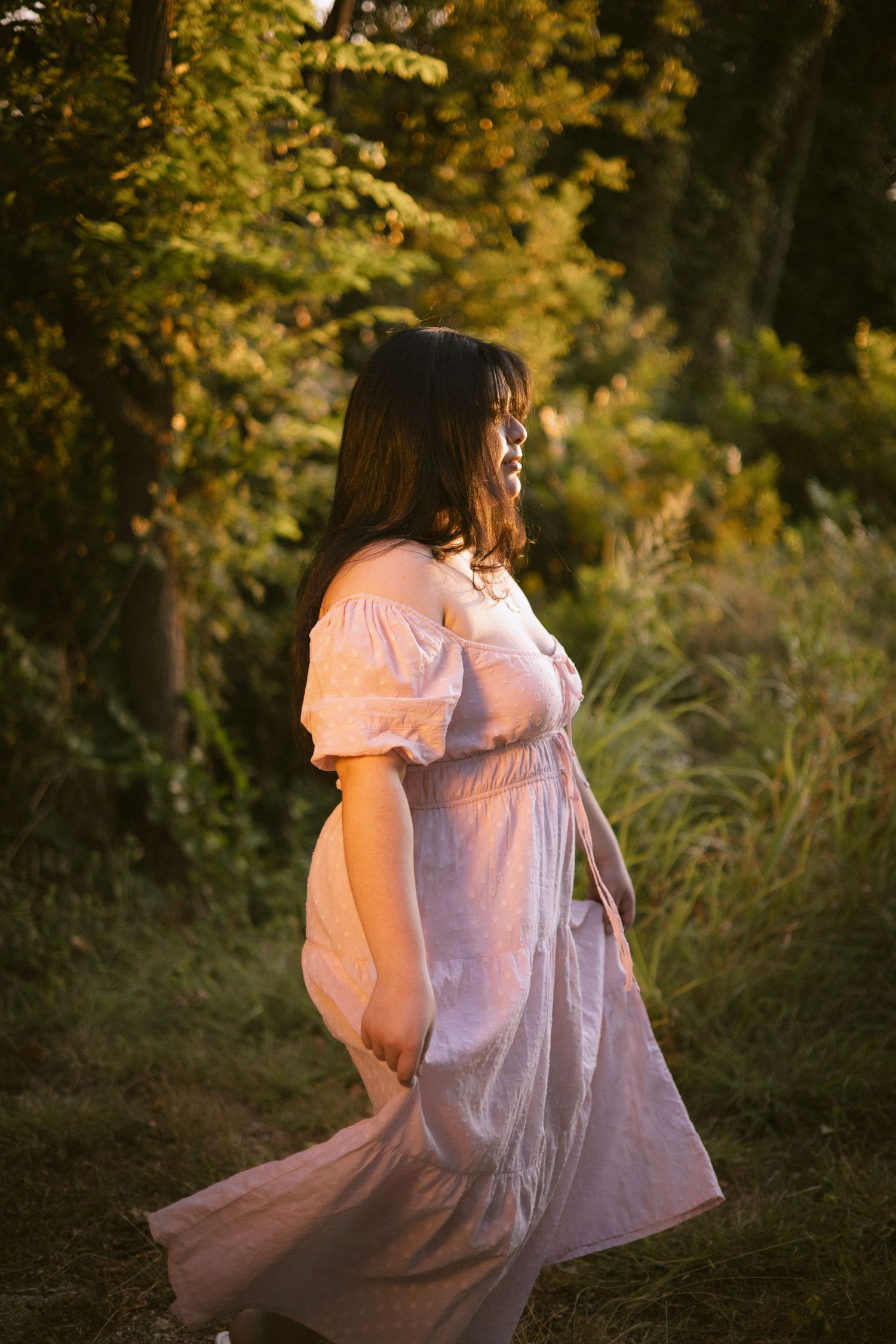 A pregnant woman in a flowing white dress stands in golden hour sunlight surrounded by lush green trees.