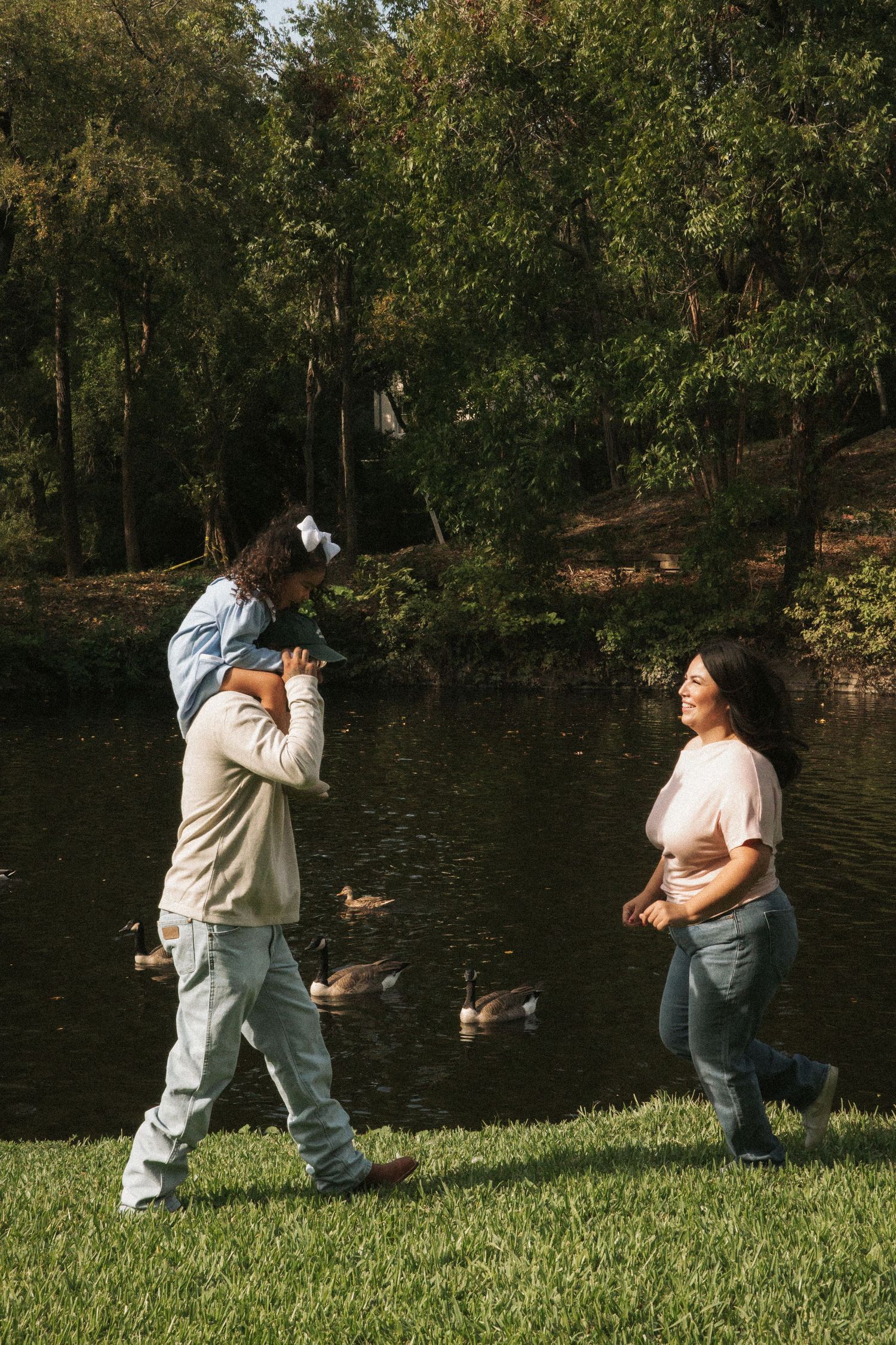 Family walking by a pond in a park, father carrying child on shoulders while mother smiles nearby.