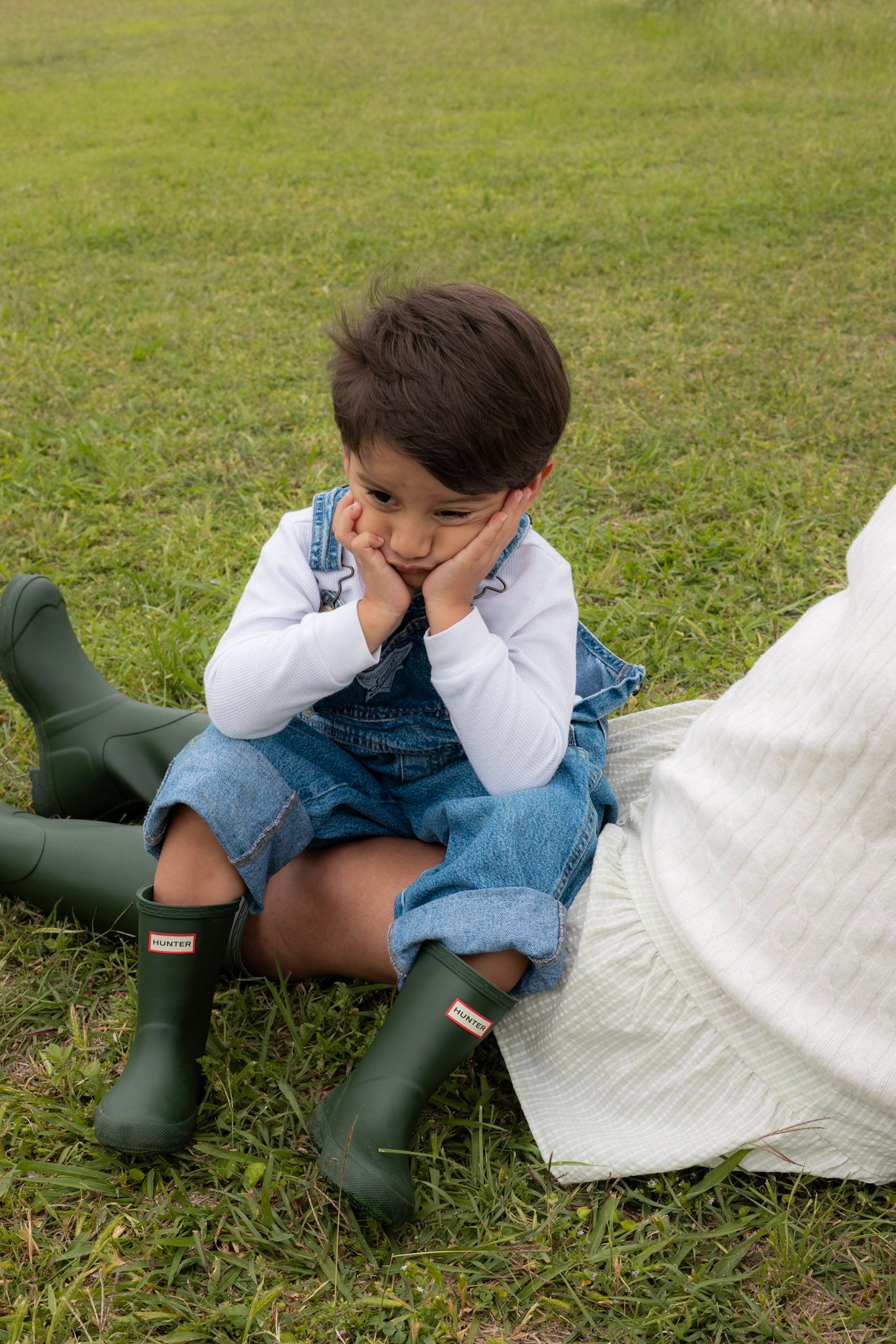 Toddler in denim overalls and green Hunter boots sitting on grass with a pouty expression.