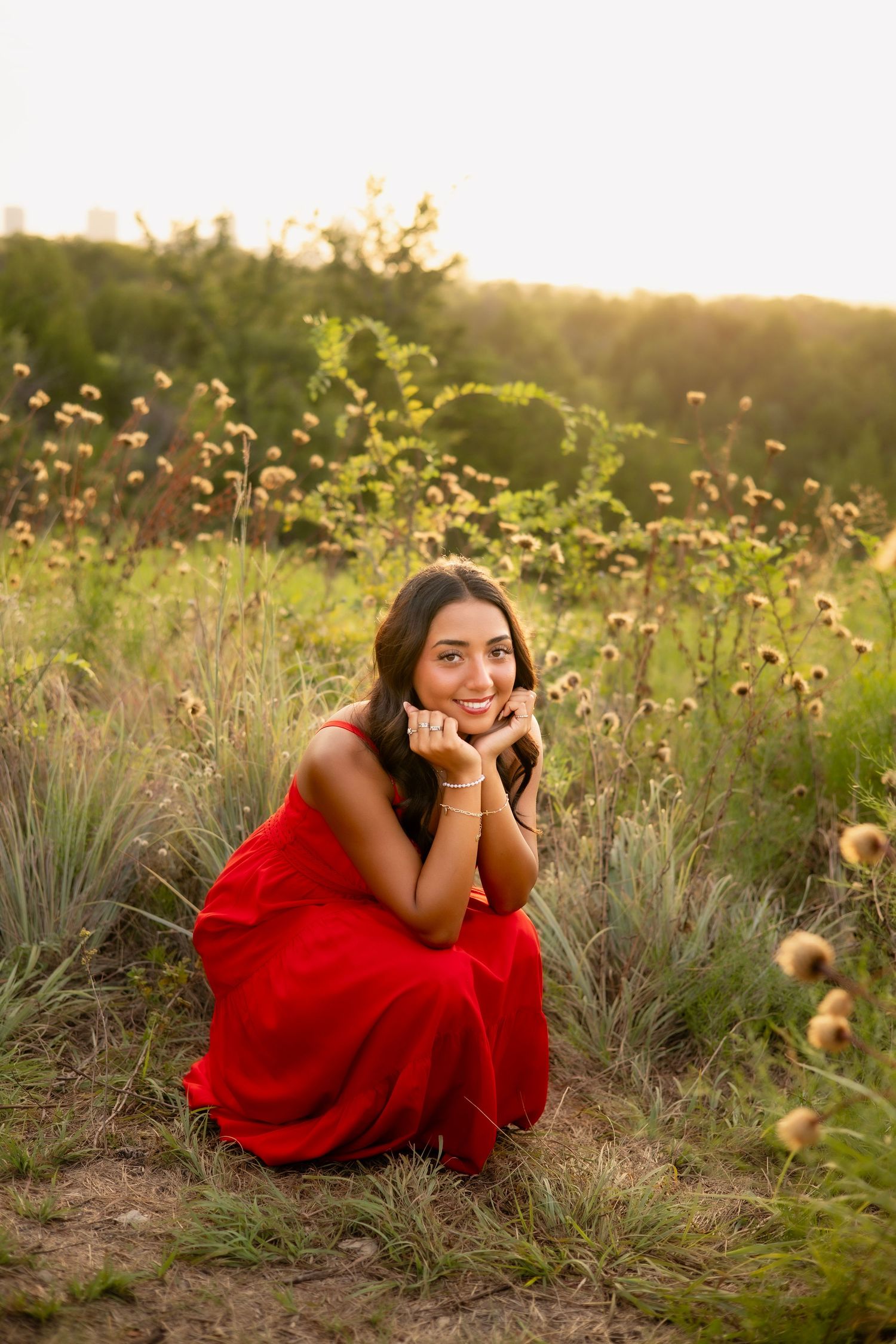 Young woman in red dress smiling at camera while seated in a wildflower field at sunset.