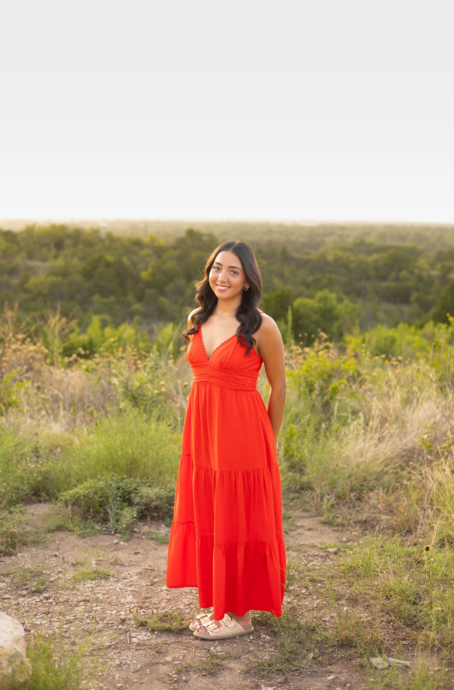 Young woman in a red maxi dress smiling outdoors in a green field during golden hour.