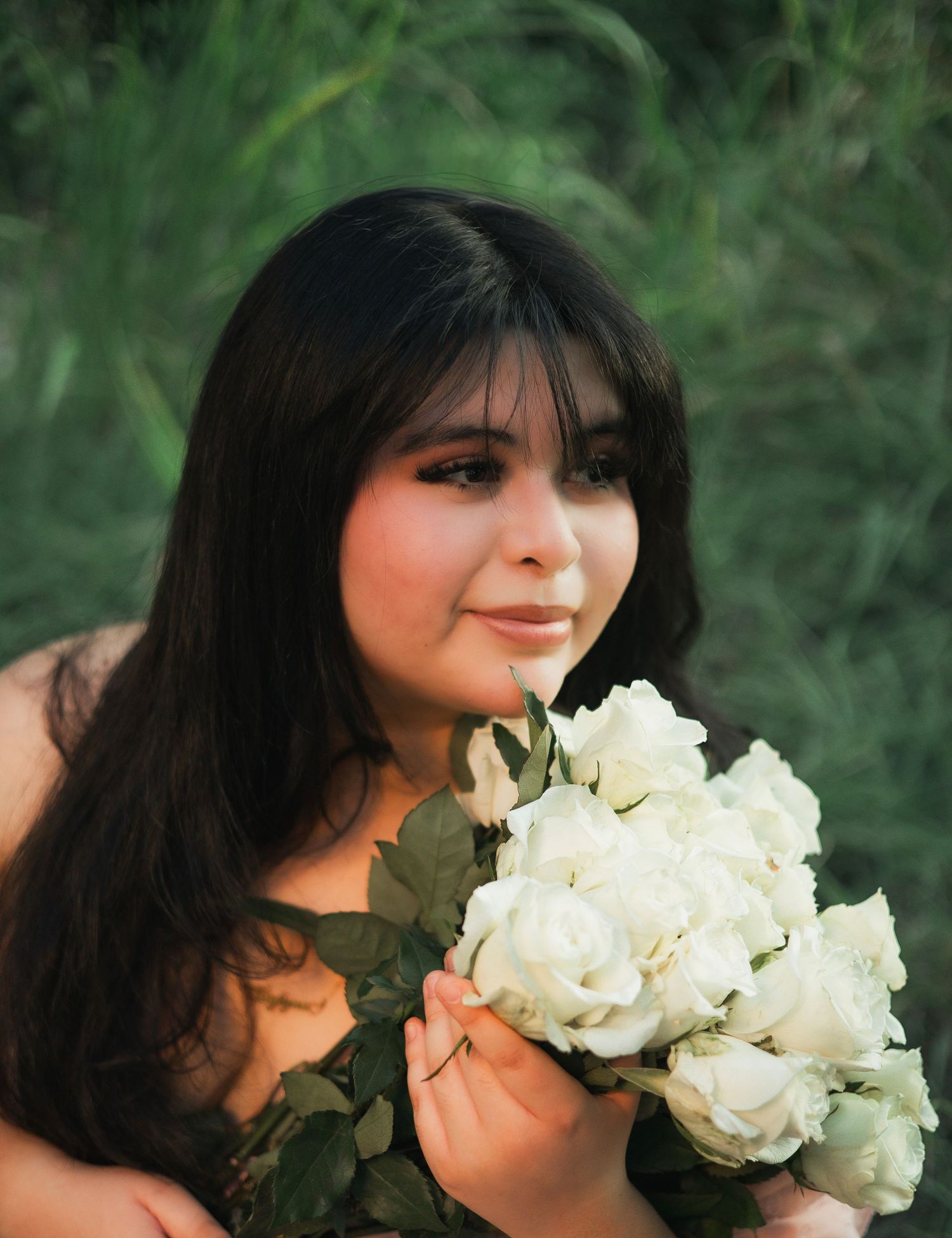 Young woman with dark hair holding white roses bouquet outdoors with green foliage background.