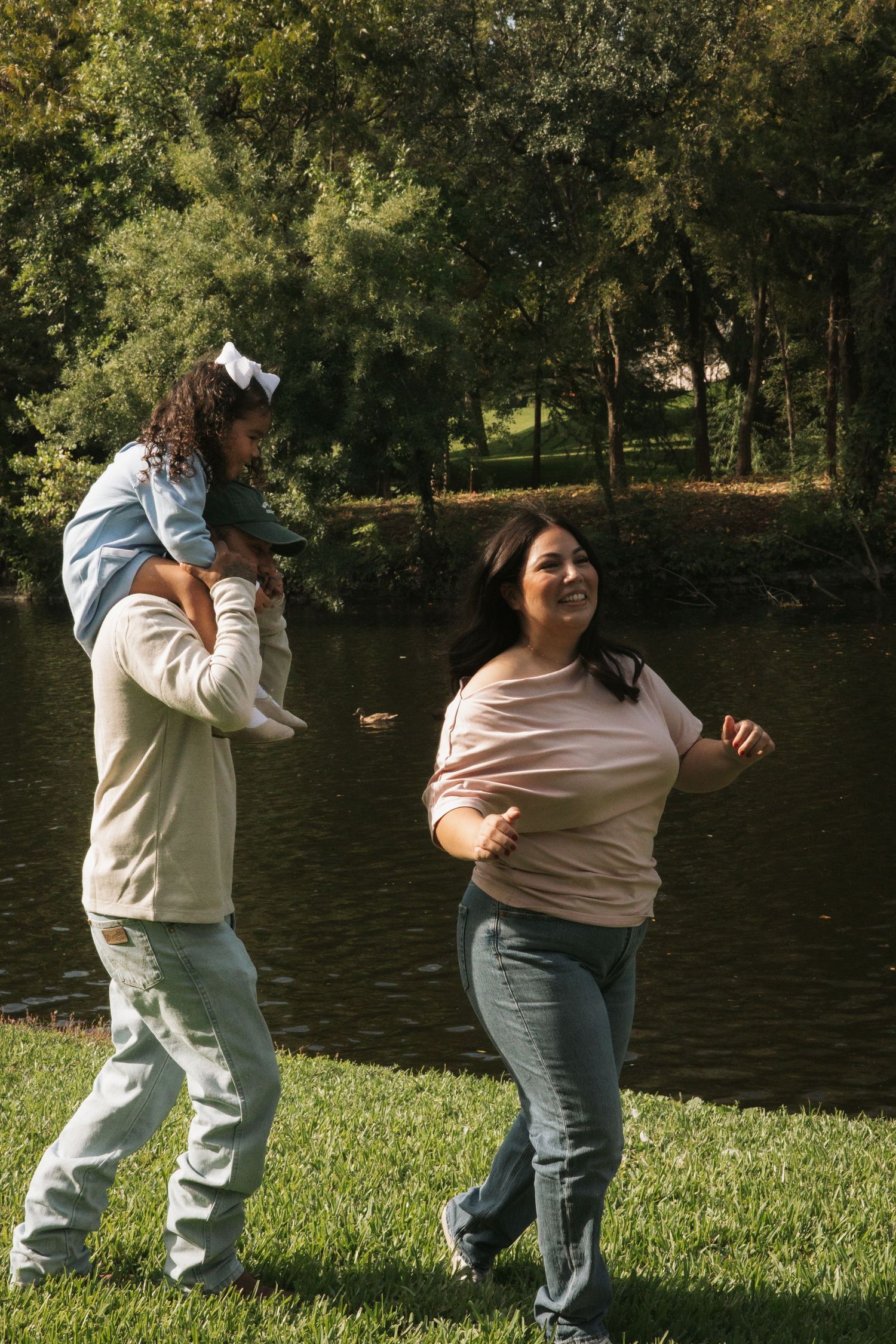 Happy family enjoying a sunny day at the park by a pond, dad carrying young girl on his shoulders.