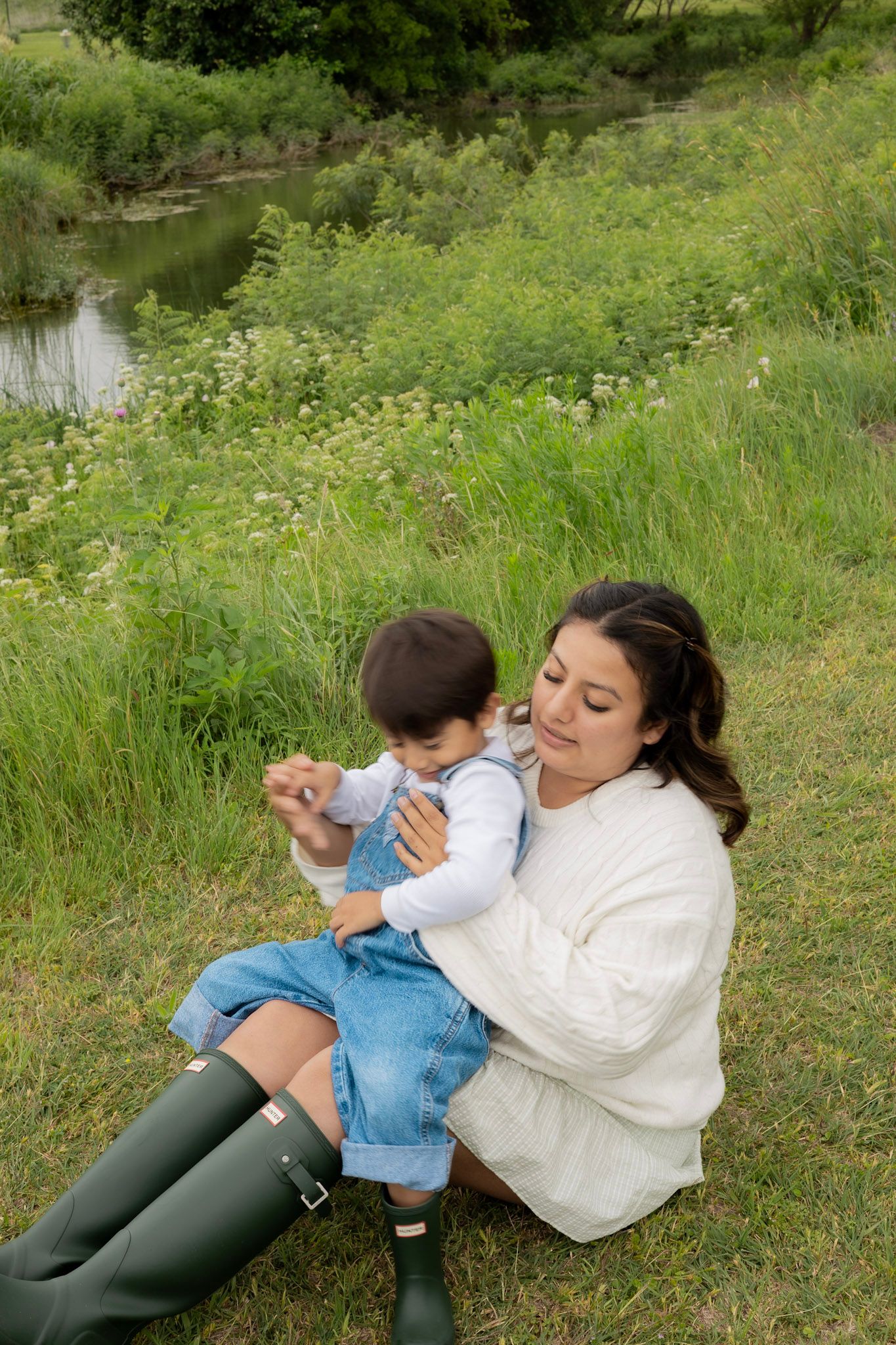 A mother sits on grass holding her toddler near a pond, wearing a white sweater and green Hunter boots.